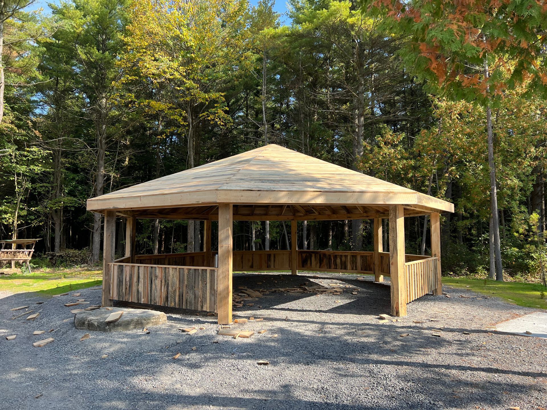 Un kiosque en bois ouvert sur les côtés, avec un toit hexagonal, se dresse sur un sol gravillonné dans une clairière.