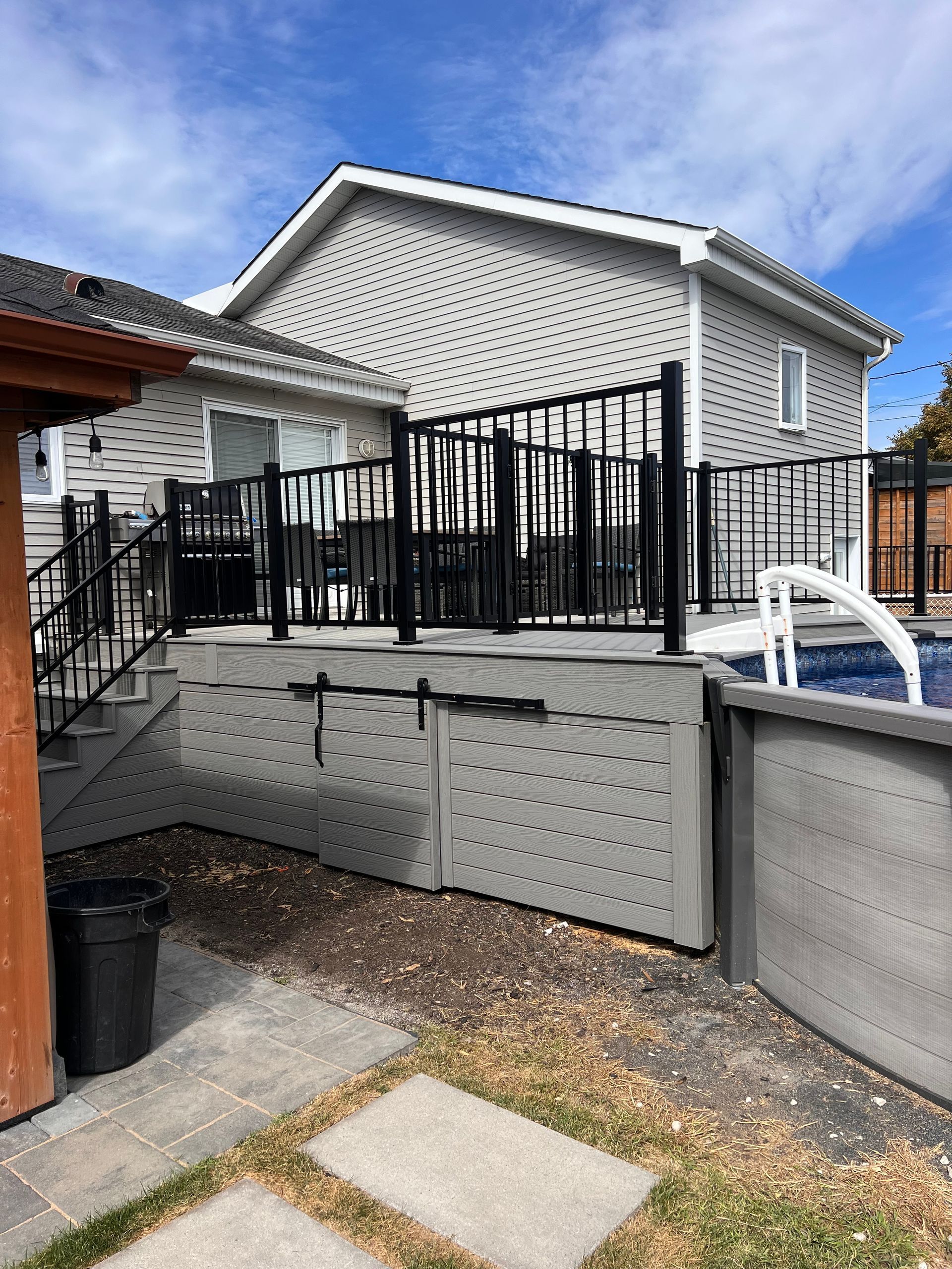 Vue d'une terrasse de maison avec des balustrades en métal noir, des plinthes en treillis gris et une piscine adjacente dans un jardin ensoleillé.