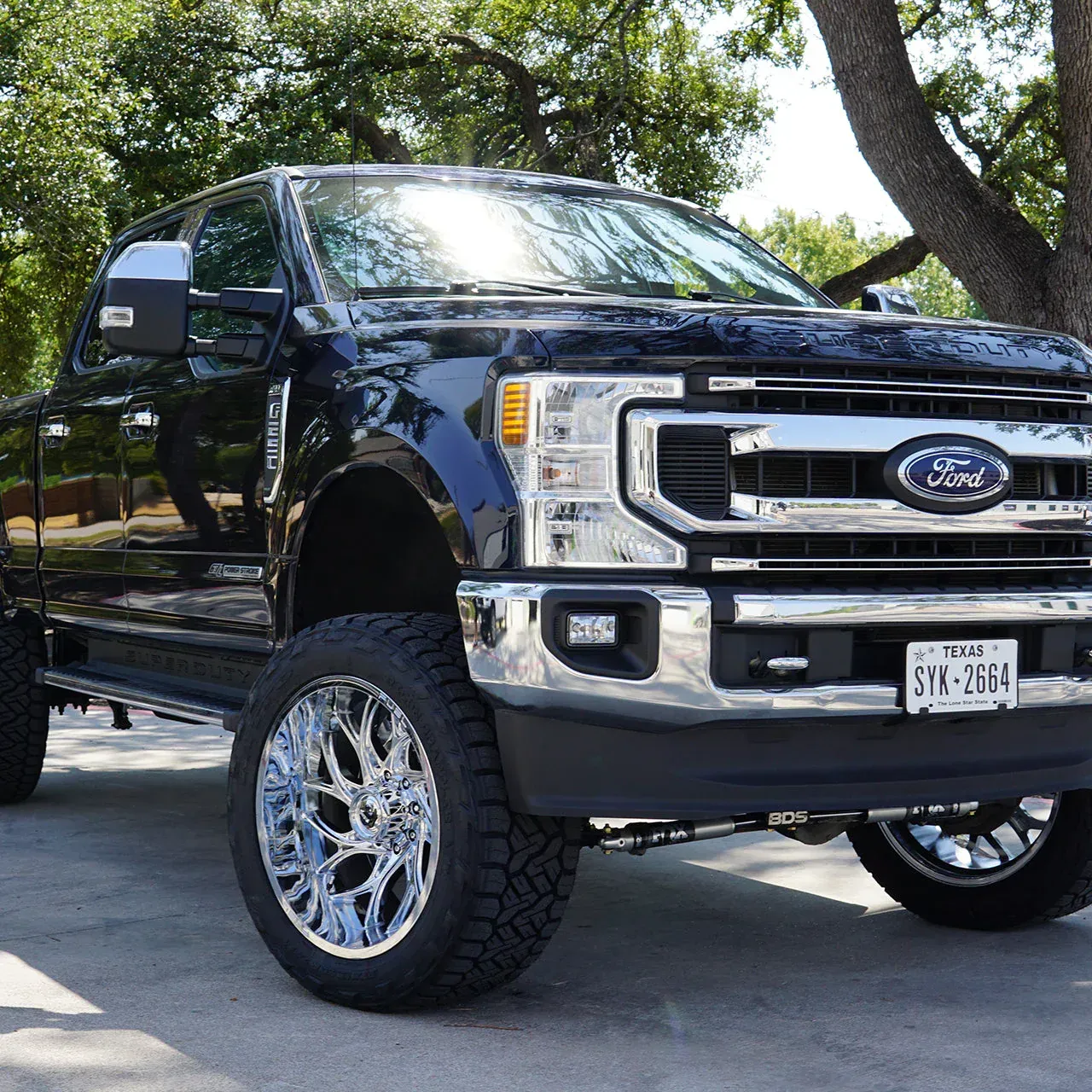 Black lifted Ford pickup truck parked outdoors on a driveway, with chrome rims and trees in the background