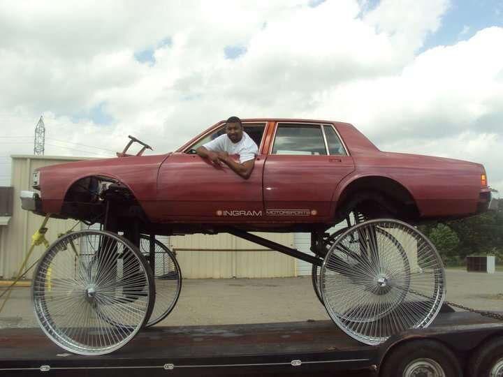 Red car body mounted on large bicycle wheels, with a person sitting inside on a flatbed trailer.