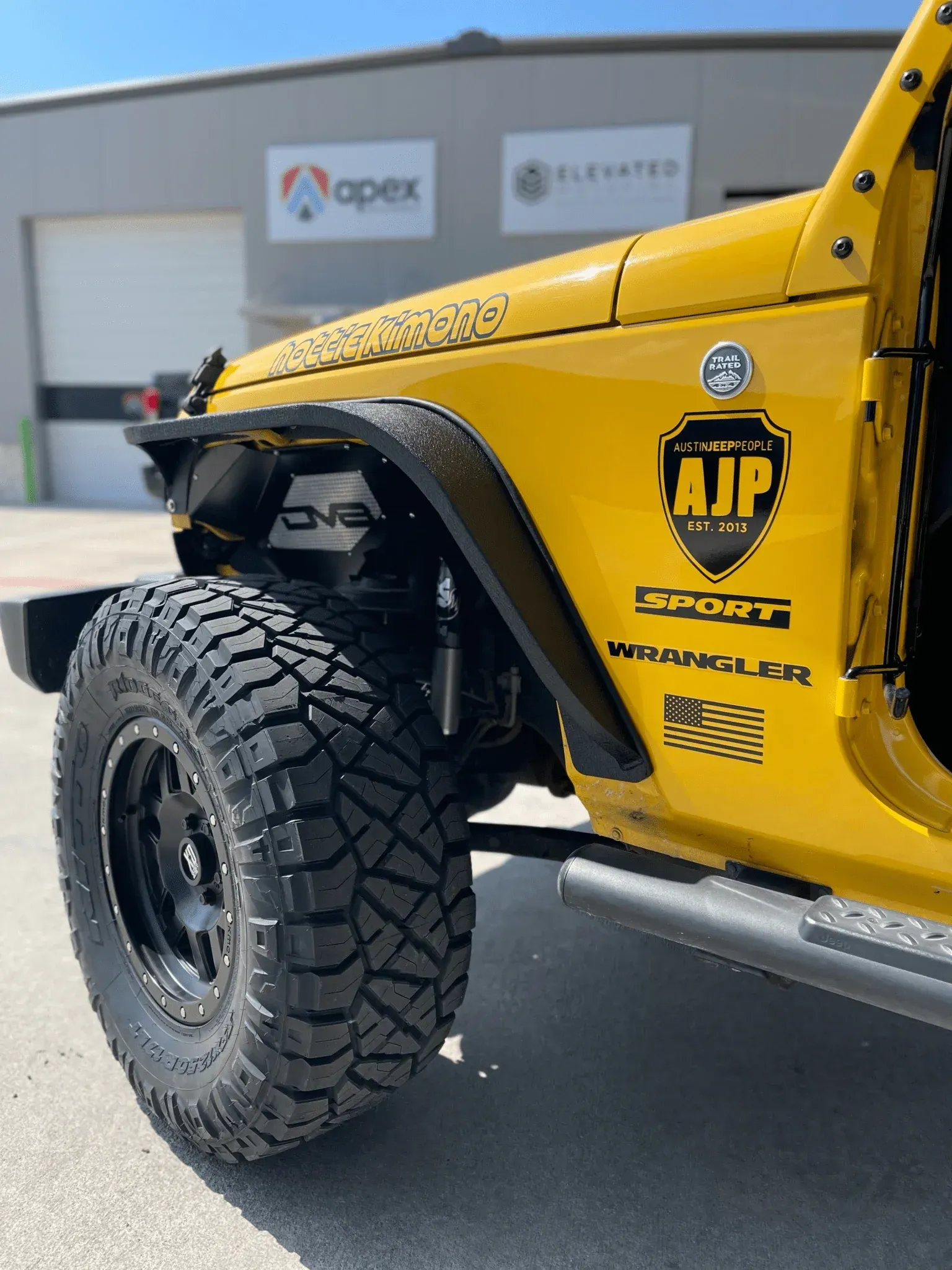 Yellow off-road Jeep with large tire and racing decals parked outside a shop