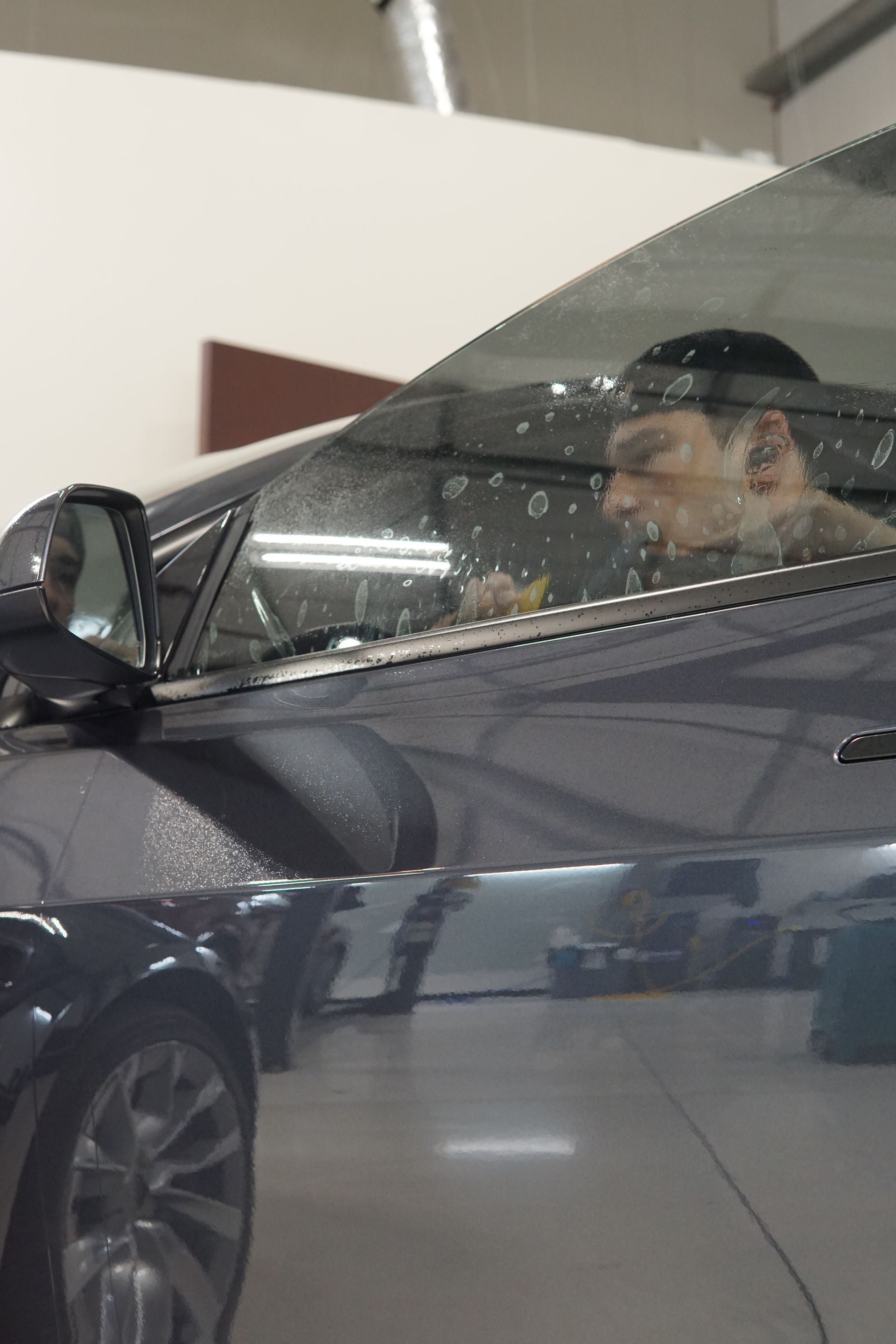 Man sitting in a dark car indoors, seen through a rain-speckled window.