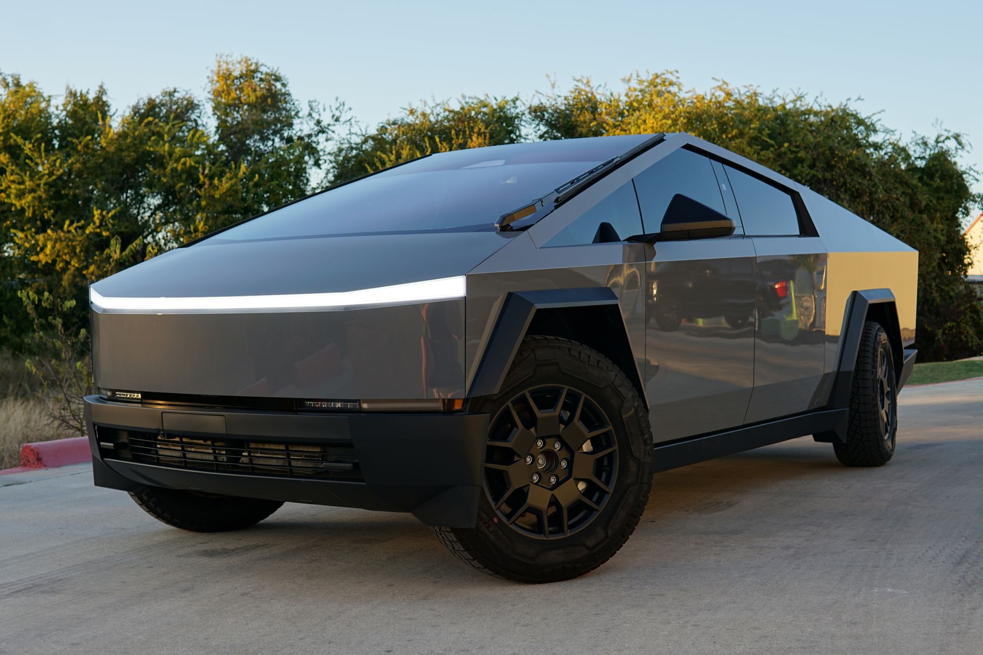 Tesla Cybertruck parked outdoors at dusk, silver angular body with illuminated front light bar.