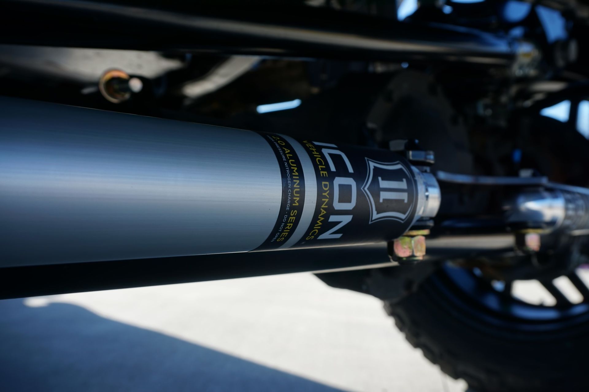 Close-up of a black suspension component beneath a vehicle, with metal fittings and a wheel in the background