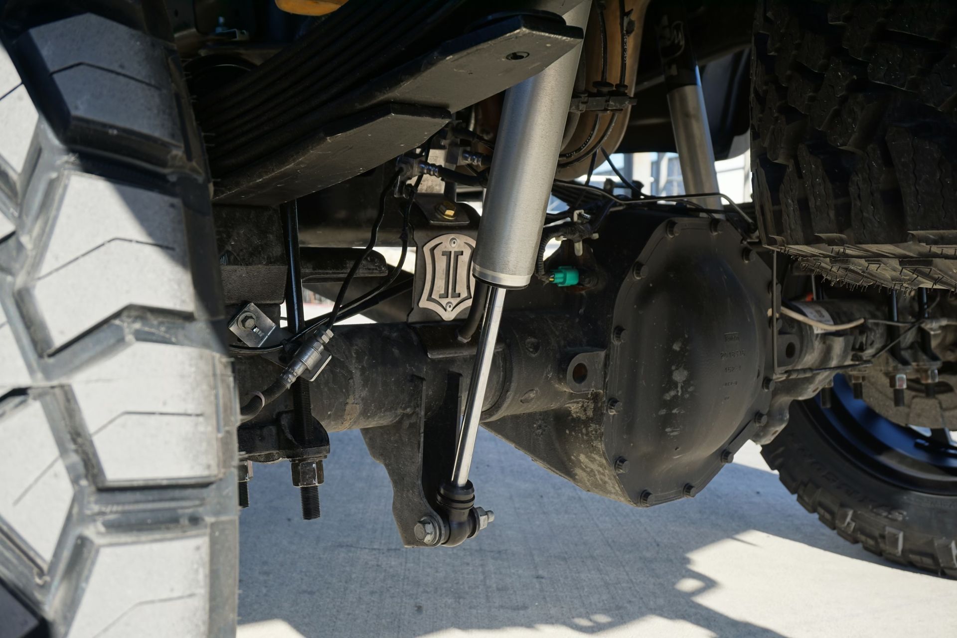 Underbody view of a truck’s suspension, axle, and large off-road tires on pavement
