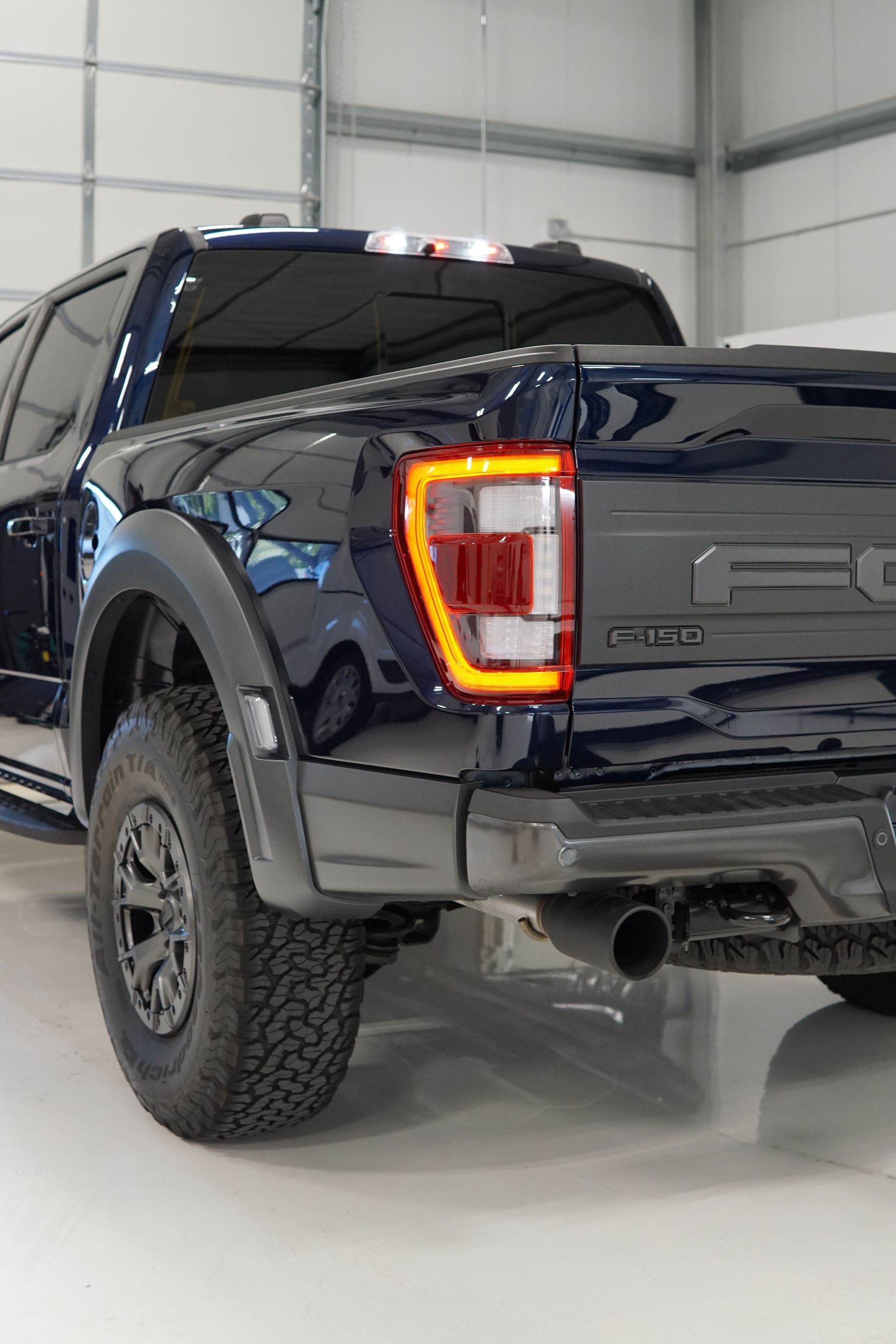 Blue Ford pickup truck rear in a white garage, with illuminated tail light and off-road tires