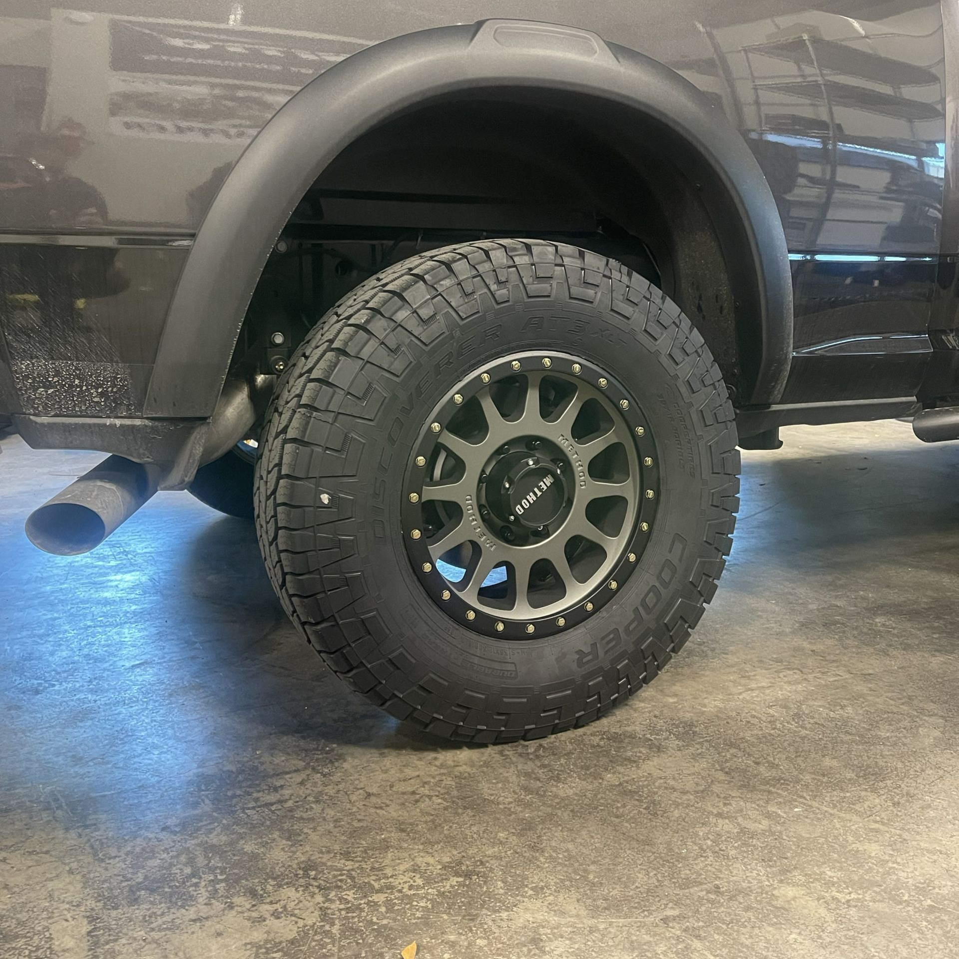 Rear wheel of a black pickup truck with off-road tire and aftermarket rim, parked on a concrete floor.