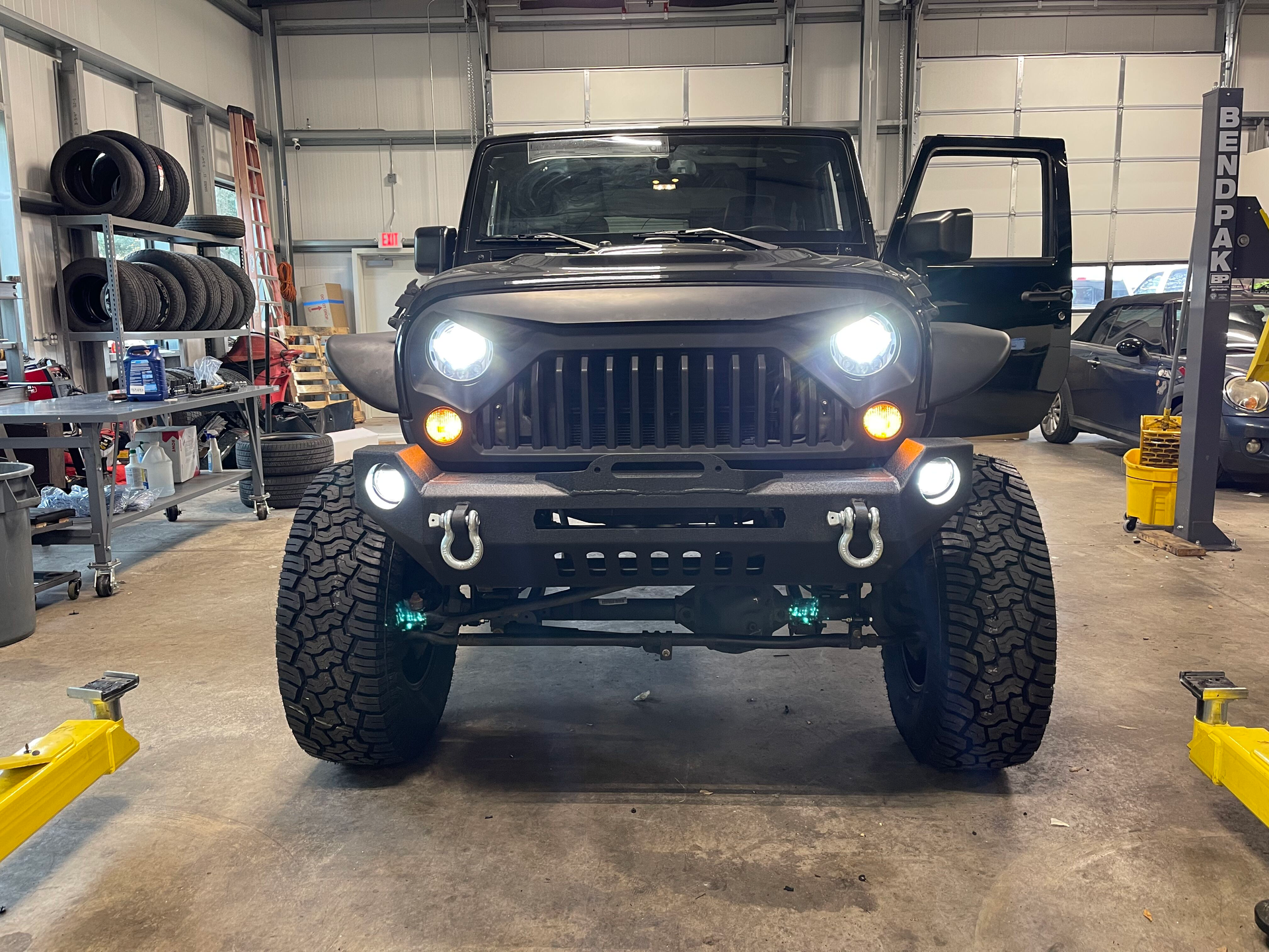 Black Jeep with headlights on in a garage, front view, lifted with off-road tires and open passenger door