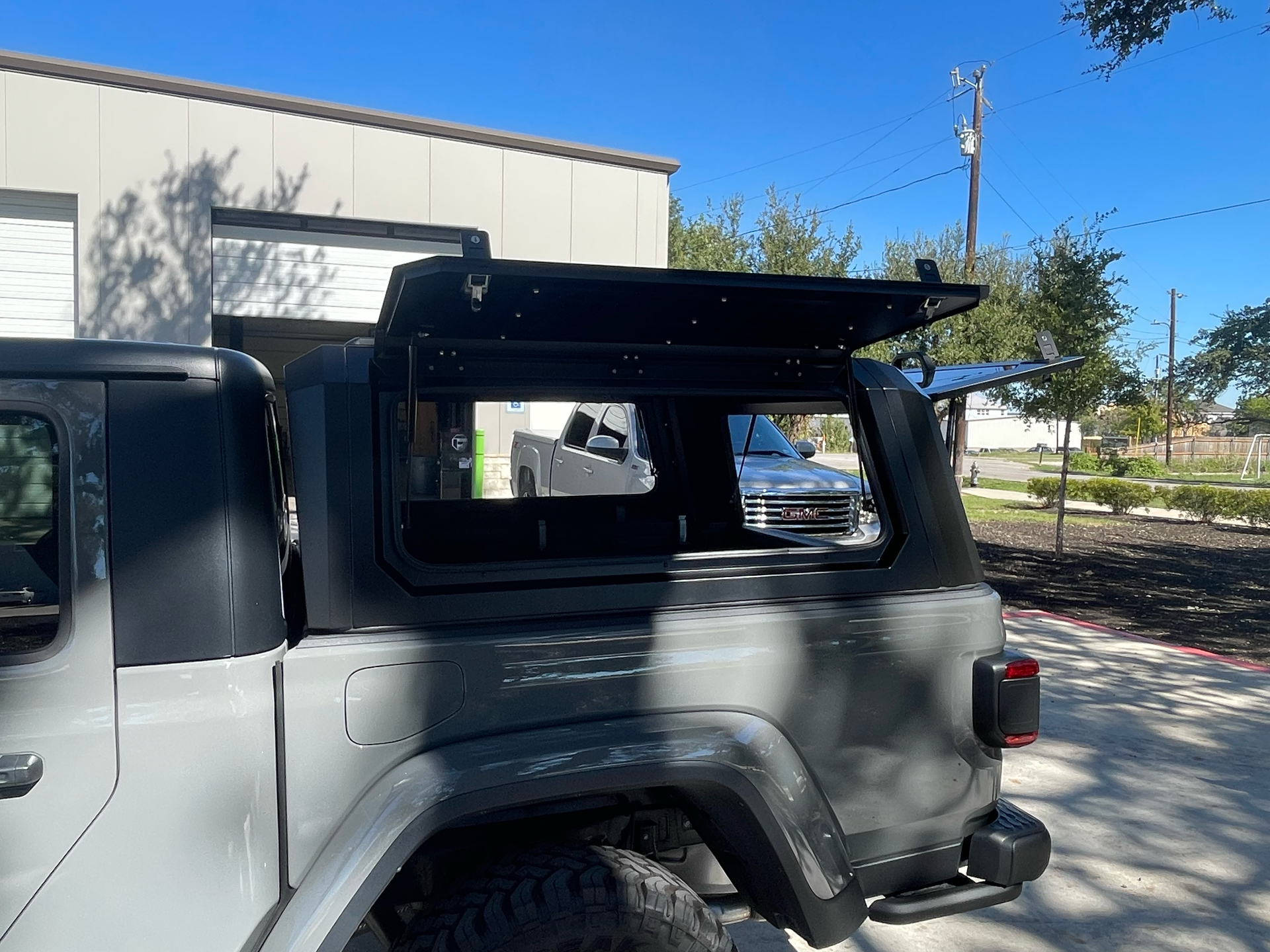 Black SUV with open rear window and roof rack parked beside a beige building on a sunny street.