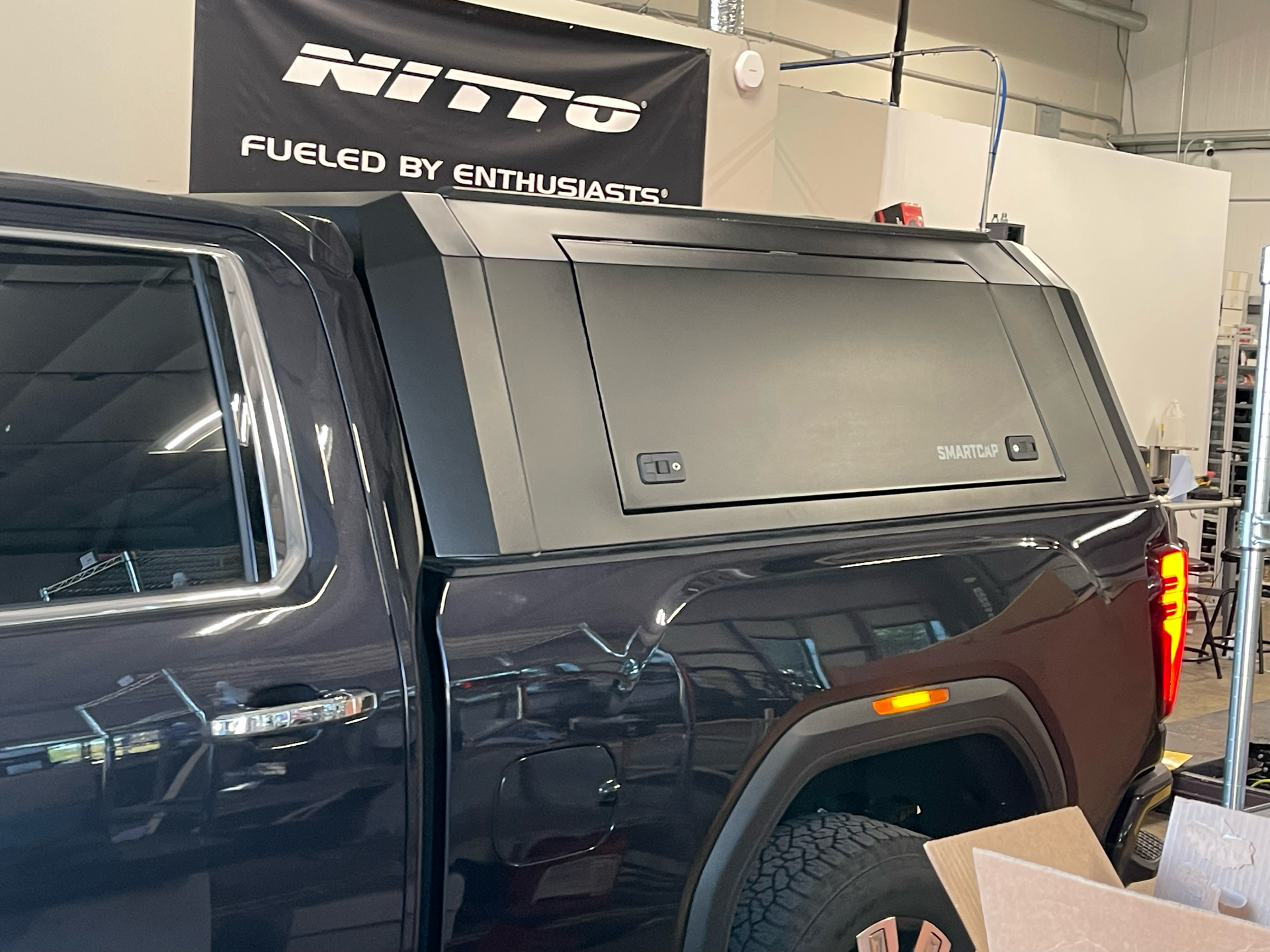 Black pickup truck with a camper shell, shown indoors near a Ritto sign.