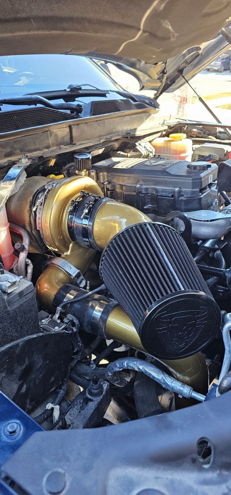 Car engine bay with a large gold turbocharger and black cone air filter under an open hood.