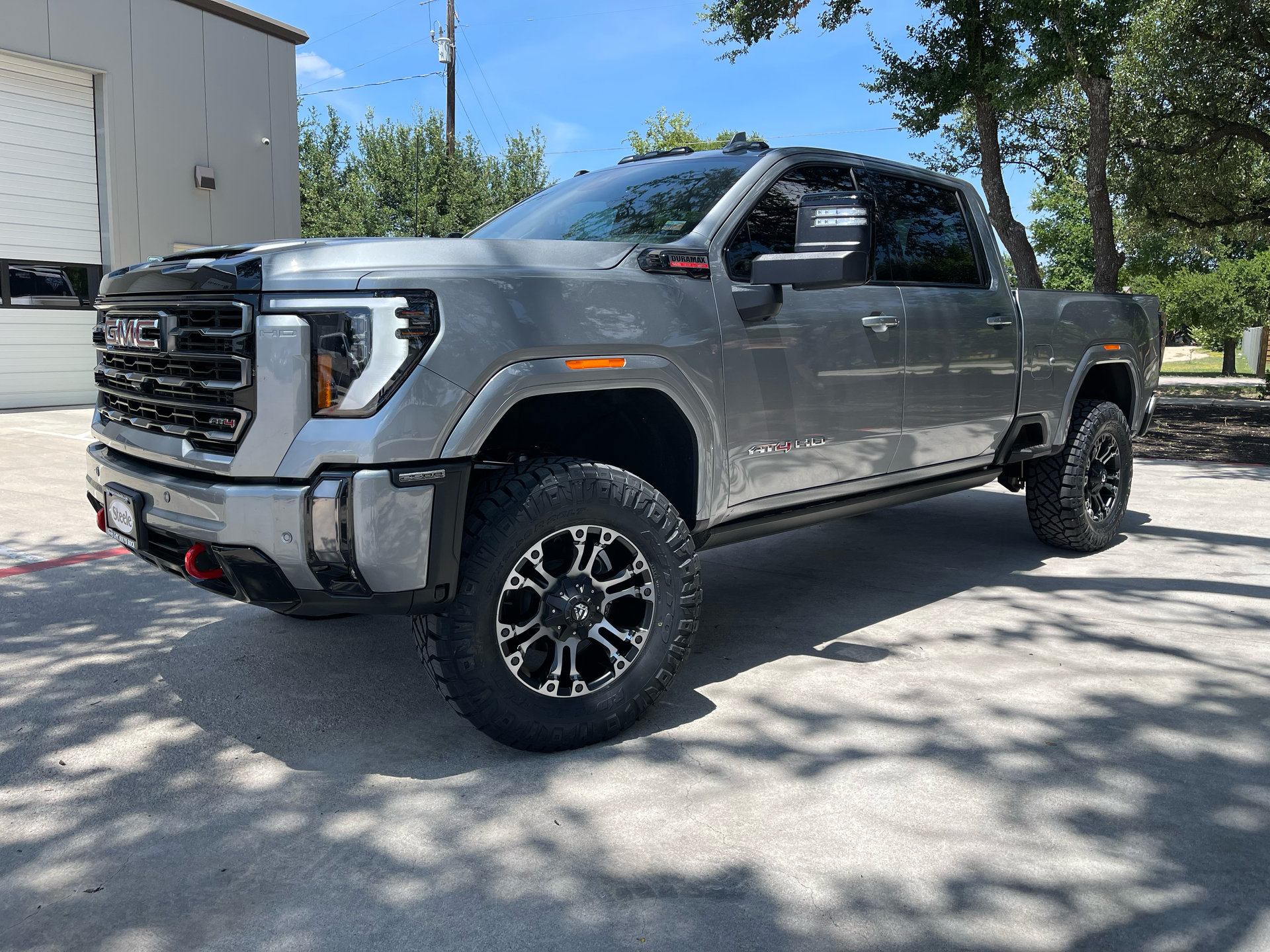 Silver lifted GMC pickup truck parked on a driveway beside a building, viewed from the front-left side
