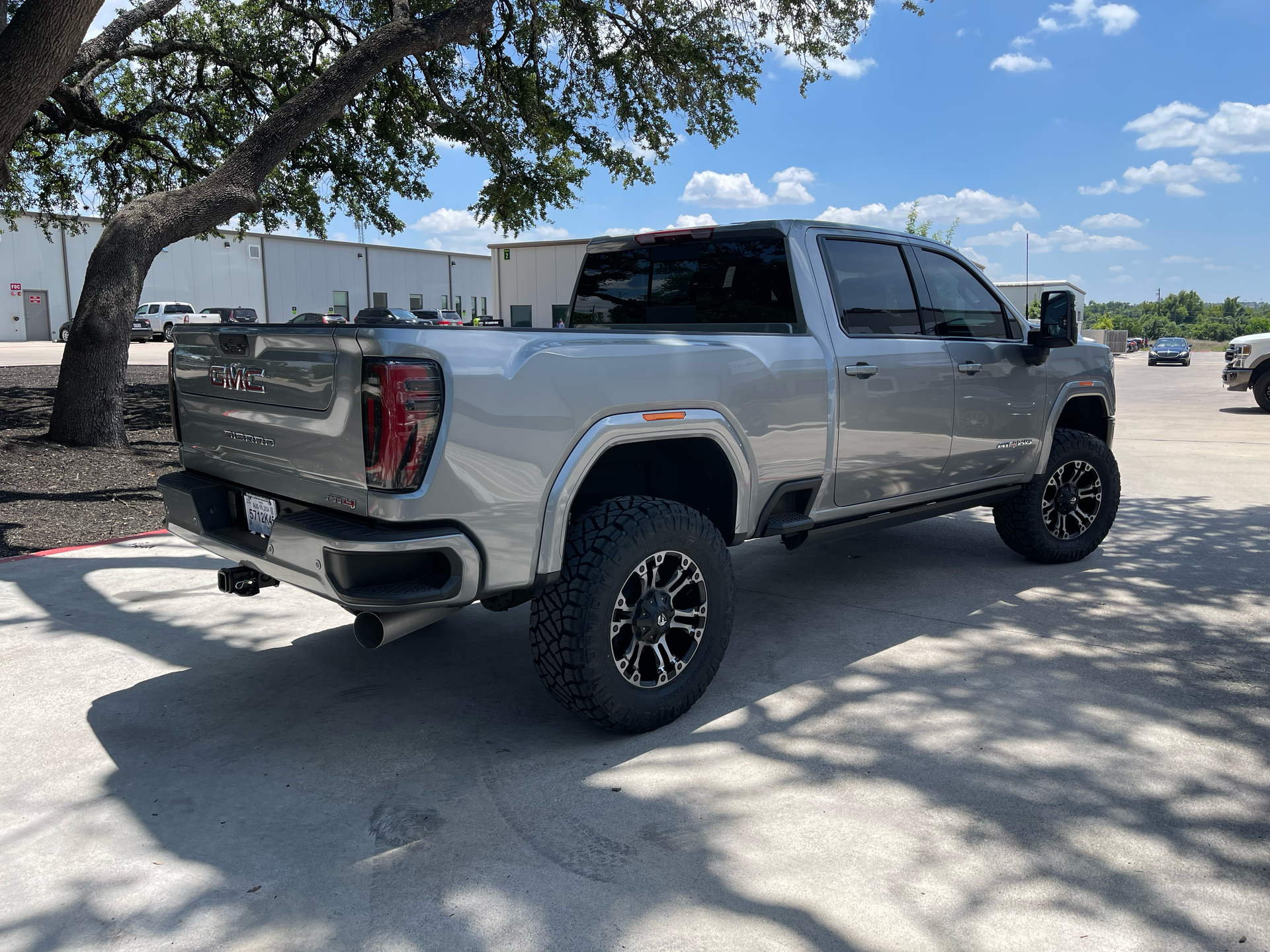 Silver lifted pickup truck parked in a sunny lot near a tree and building