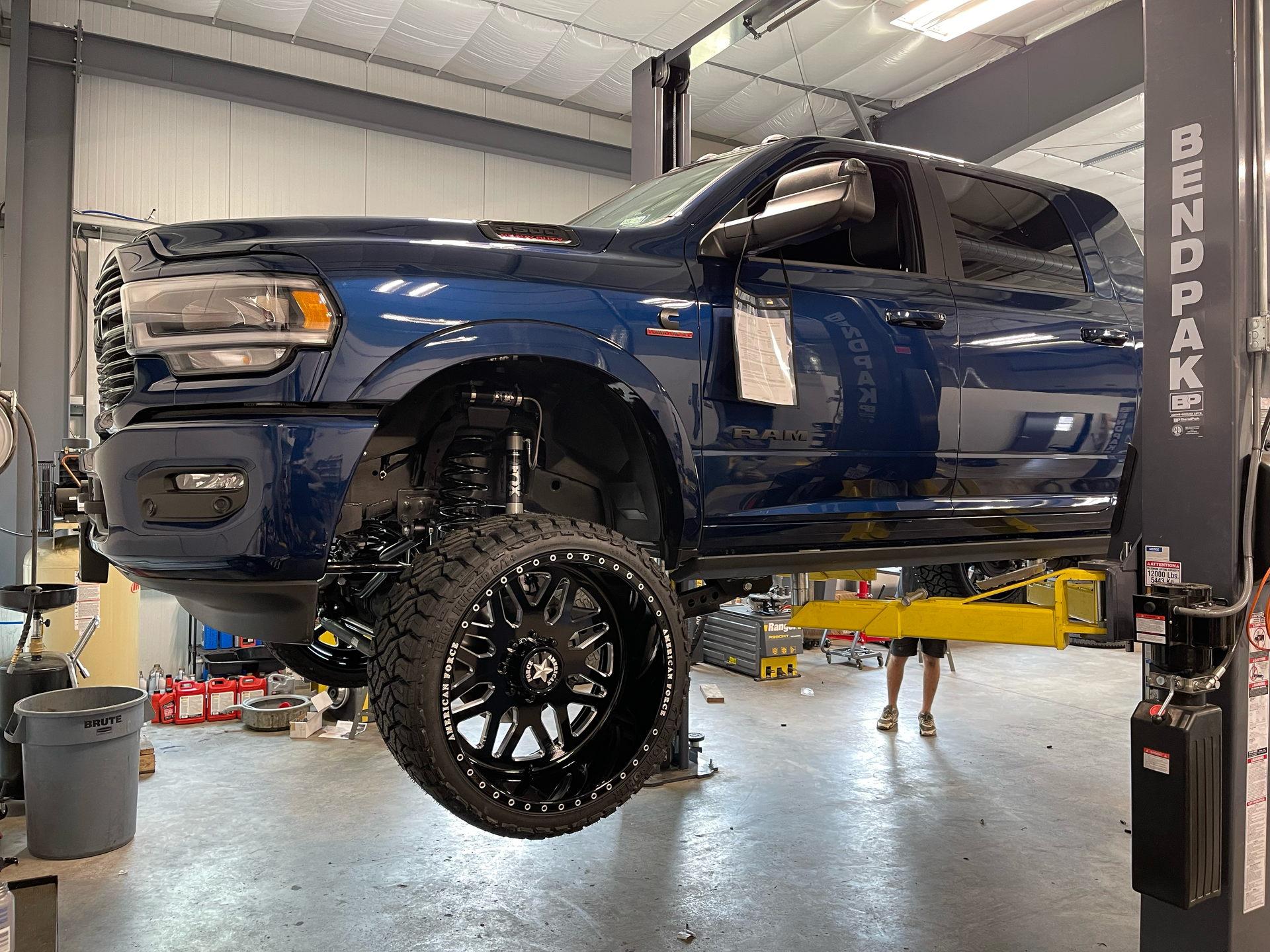 Lifted blue pickup truck in an auto shop on a hydraulic lift, with a wheel removed.