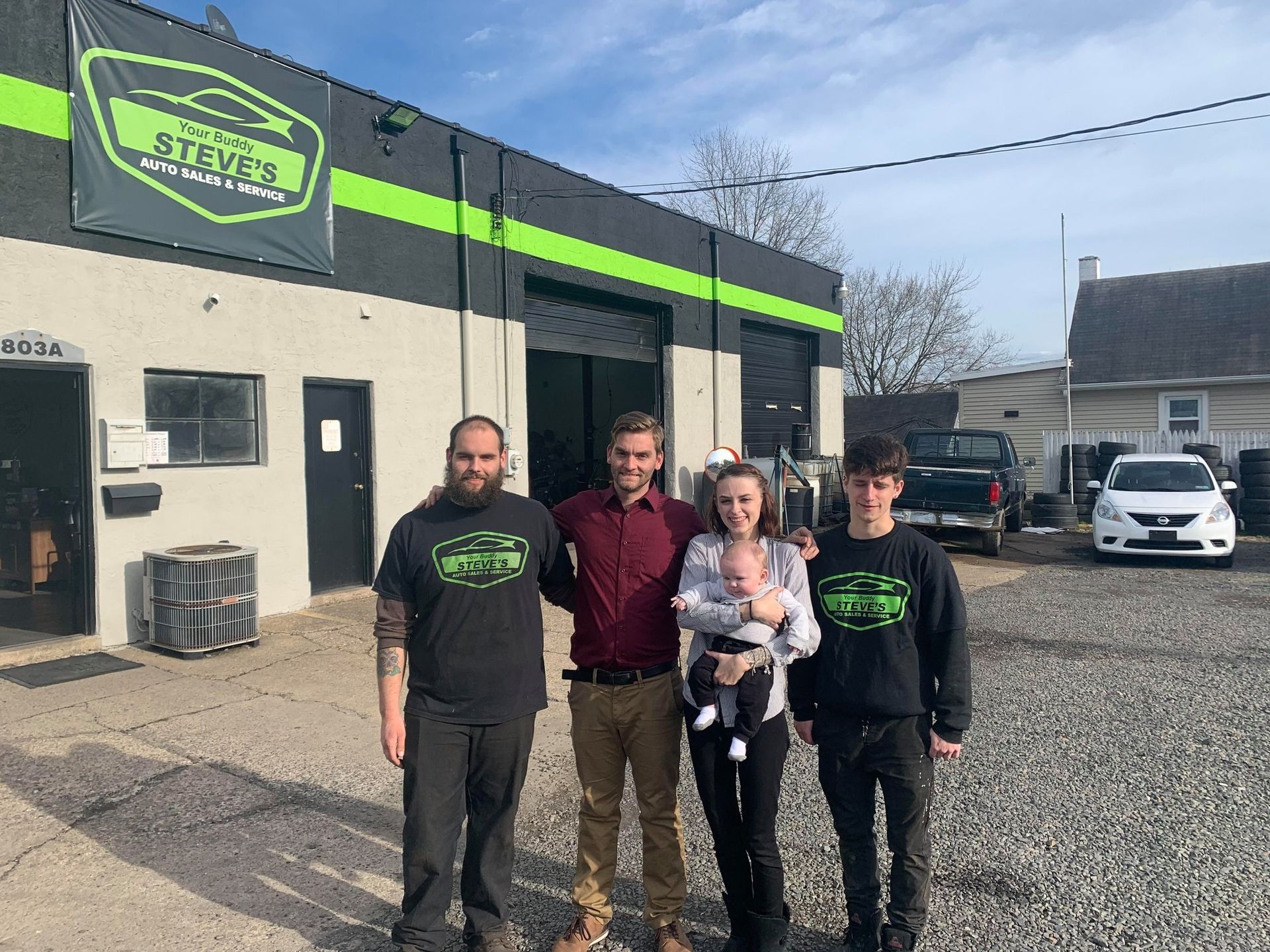 Four people standing outside Steve's Automotive garage; smiling. One person holds a baby. The building is gray and black. | Your Buddy Steve's Garage