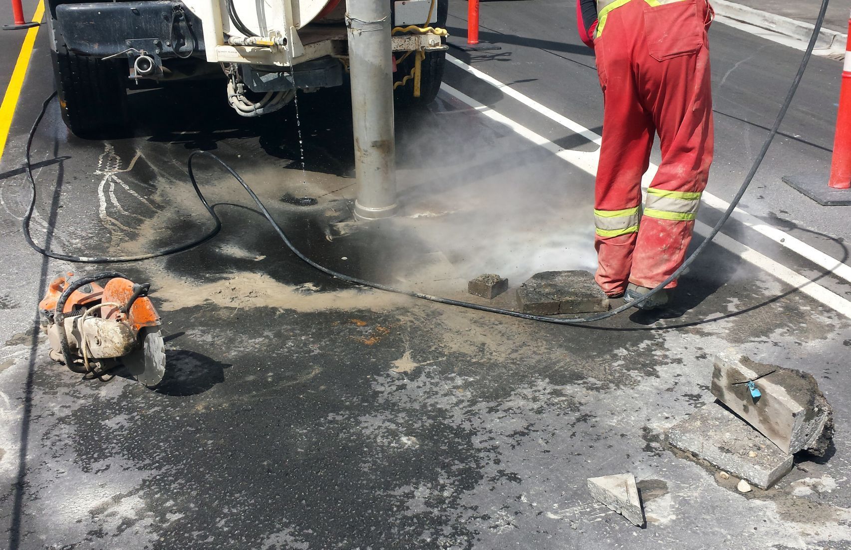 Equipe de obras reparando o pavimento ao redor de um poste de energia com um martelo pneumático e outras ferramentas, em meio à poeira e detritos.