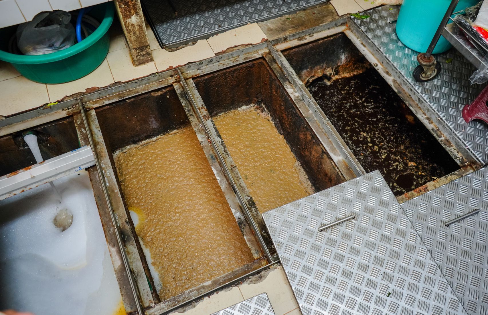Painéis de acesso ao piso aberto revelam compartimentos escuros e líquido amarelo em um espaço industrial.