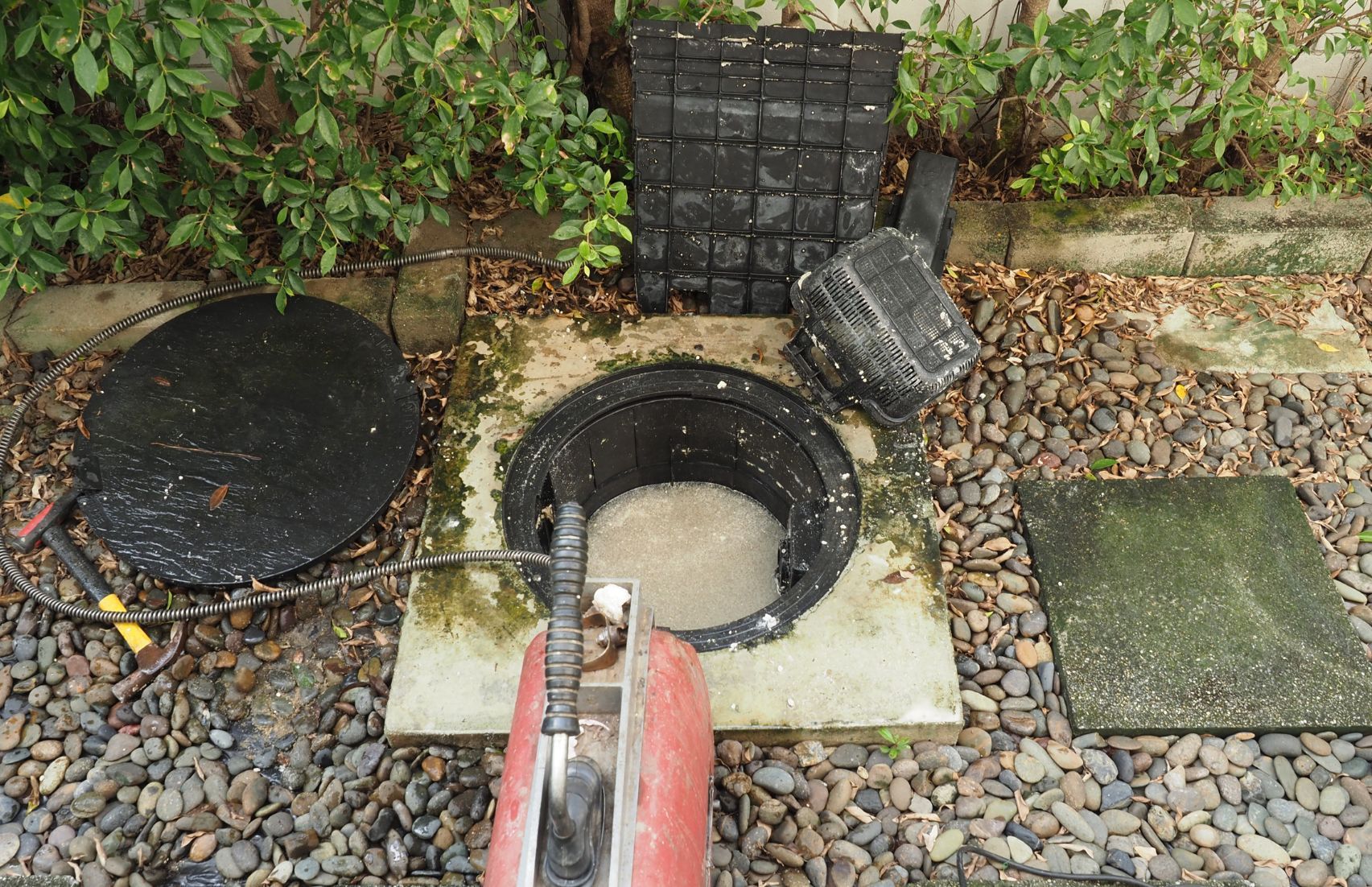 Bueiro redondo aberto com água, ao lado de tampas de serviços públicos pretas e caminho de jardim com pedras.