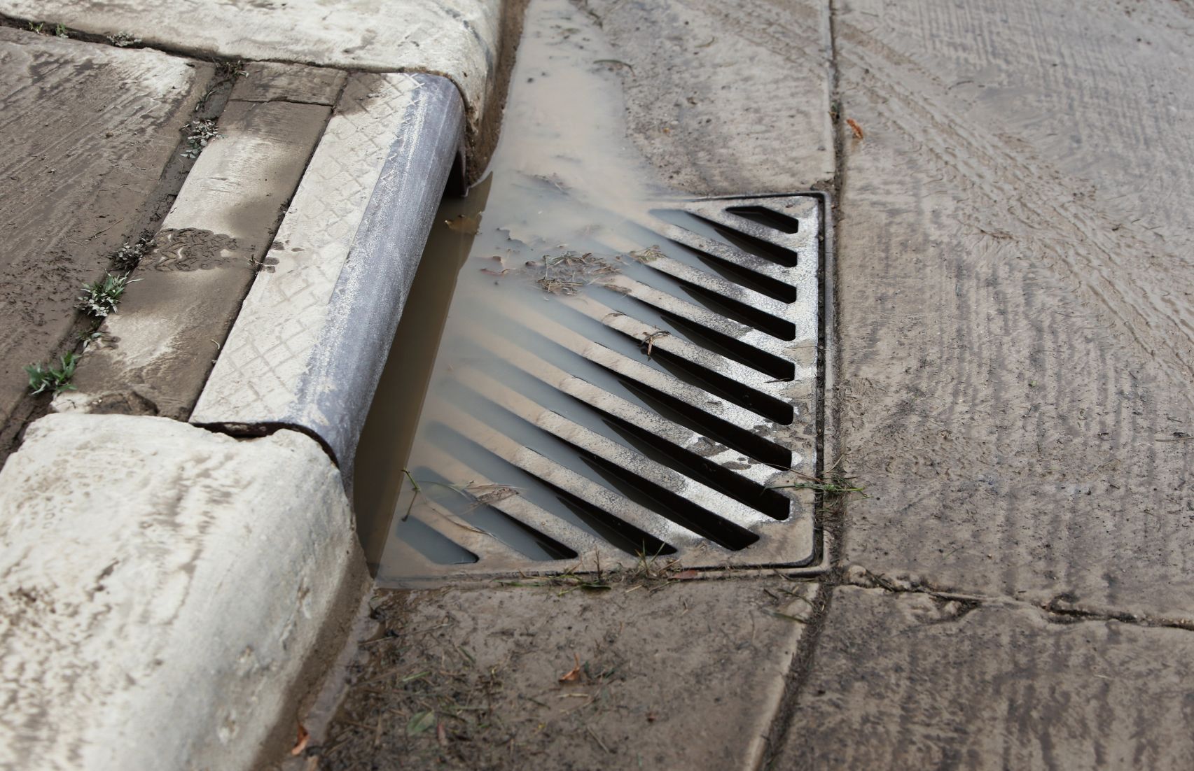 Grelha de drenagem pluvial ao lado de um meio-fio em uma rua de concreto, com água escorrendo pela borda.