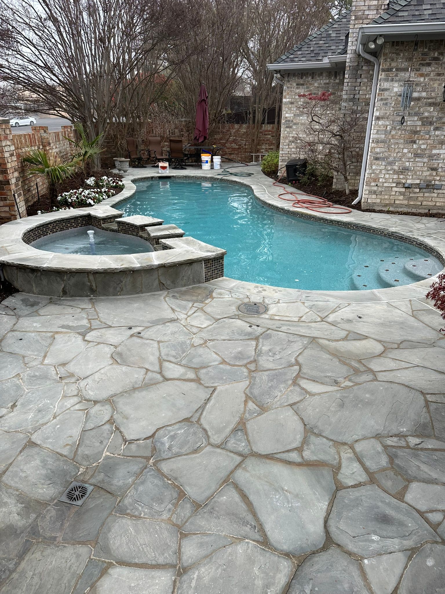 A backyard with a pool and spa surrounded by stone patio. Buildings in the background.
