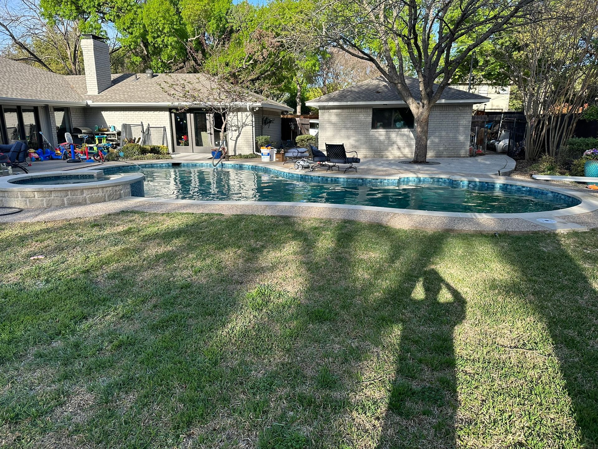 Backyard with a swimming pool and spa. Green lawn, trees, a stone pool deck, and a small building are visible.