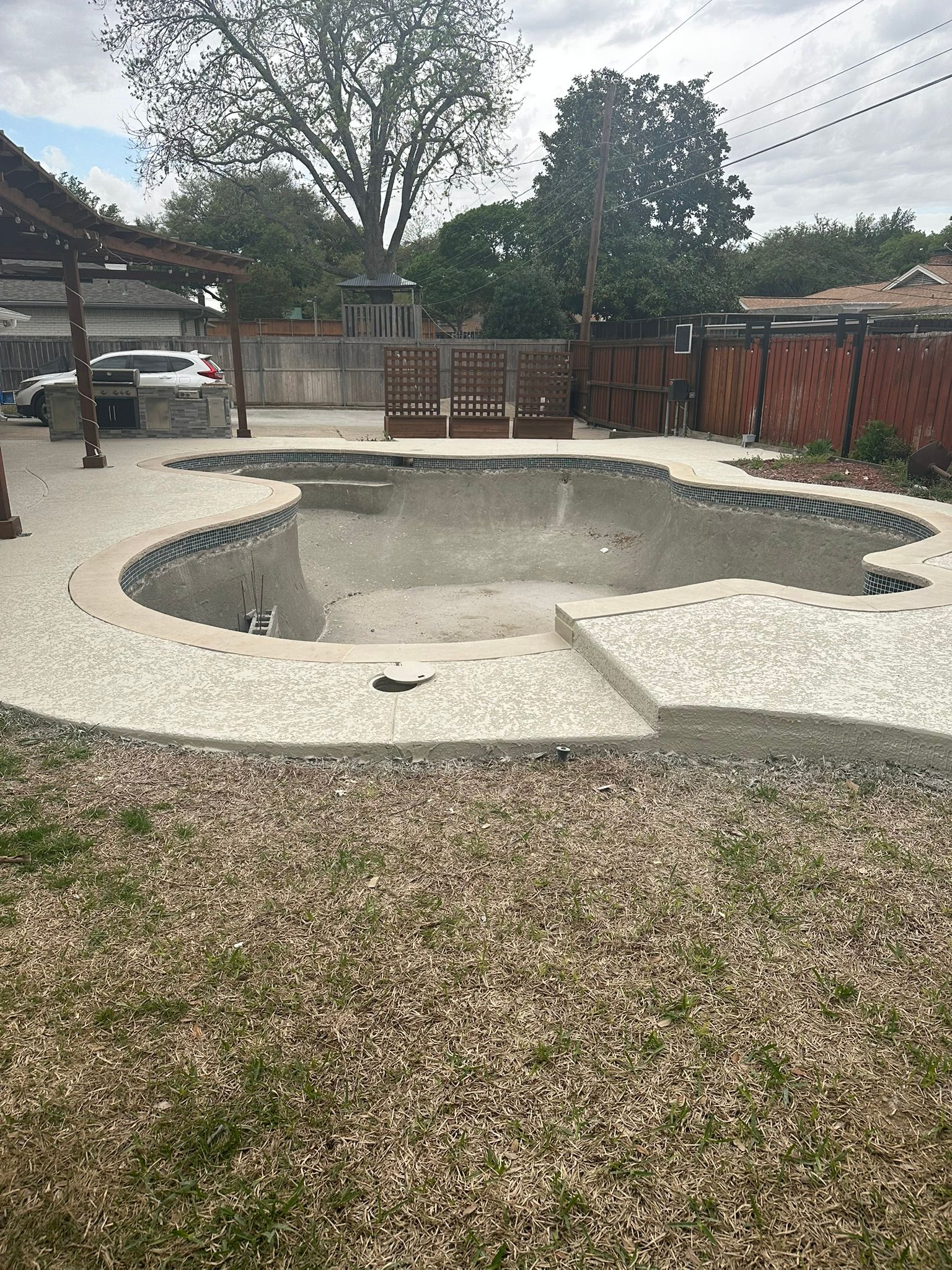 Empty swimming pool in a backyard, surrounded by gravel and a wooden fence. Overcast sky.