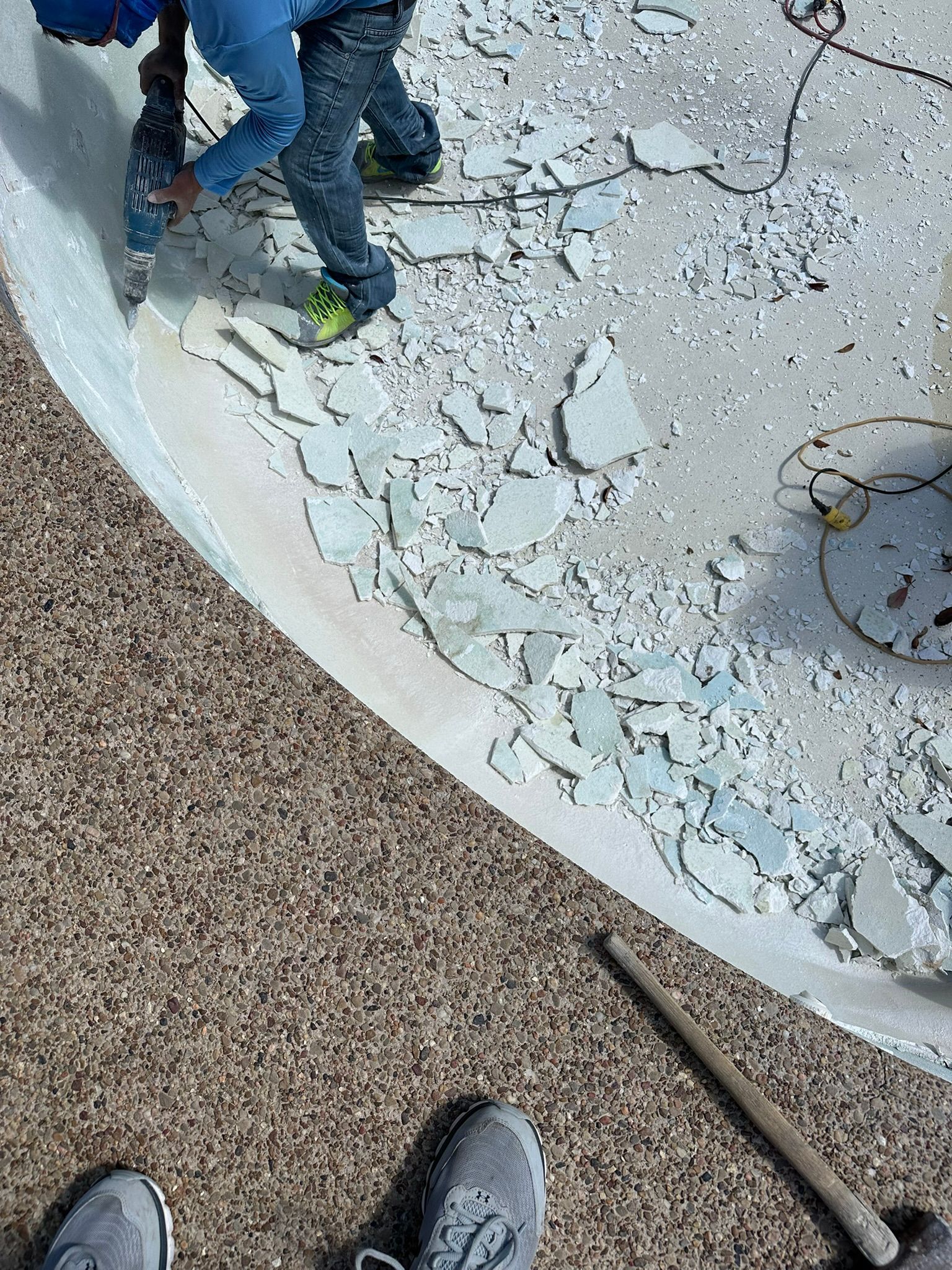 Person using a jackhammer to break up a pool's tile border; debris scattered.