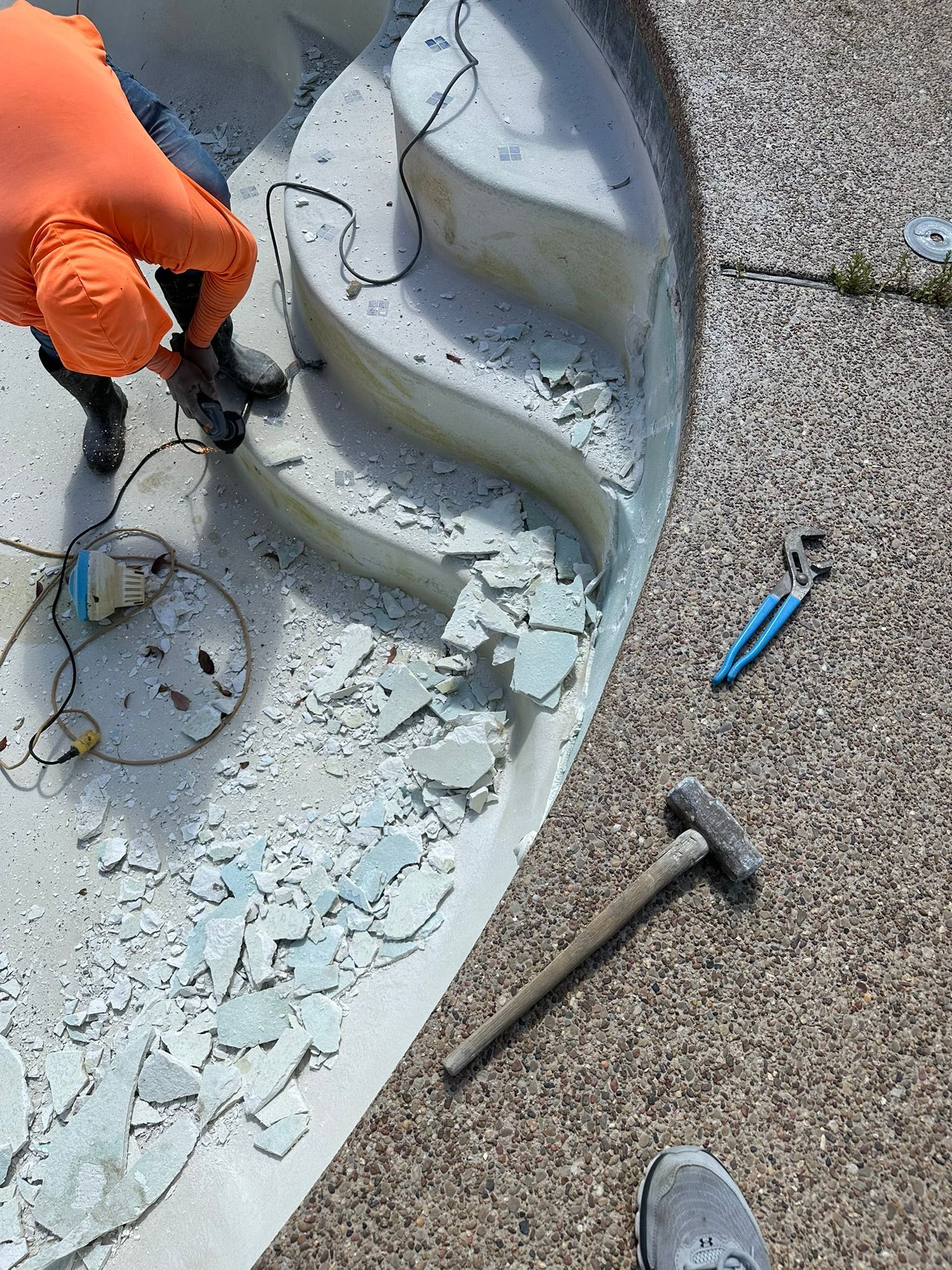 Person chipping away at a pool's tile with tools. Orange gloved hand, hammer, and pliers visible.