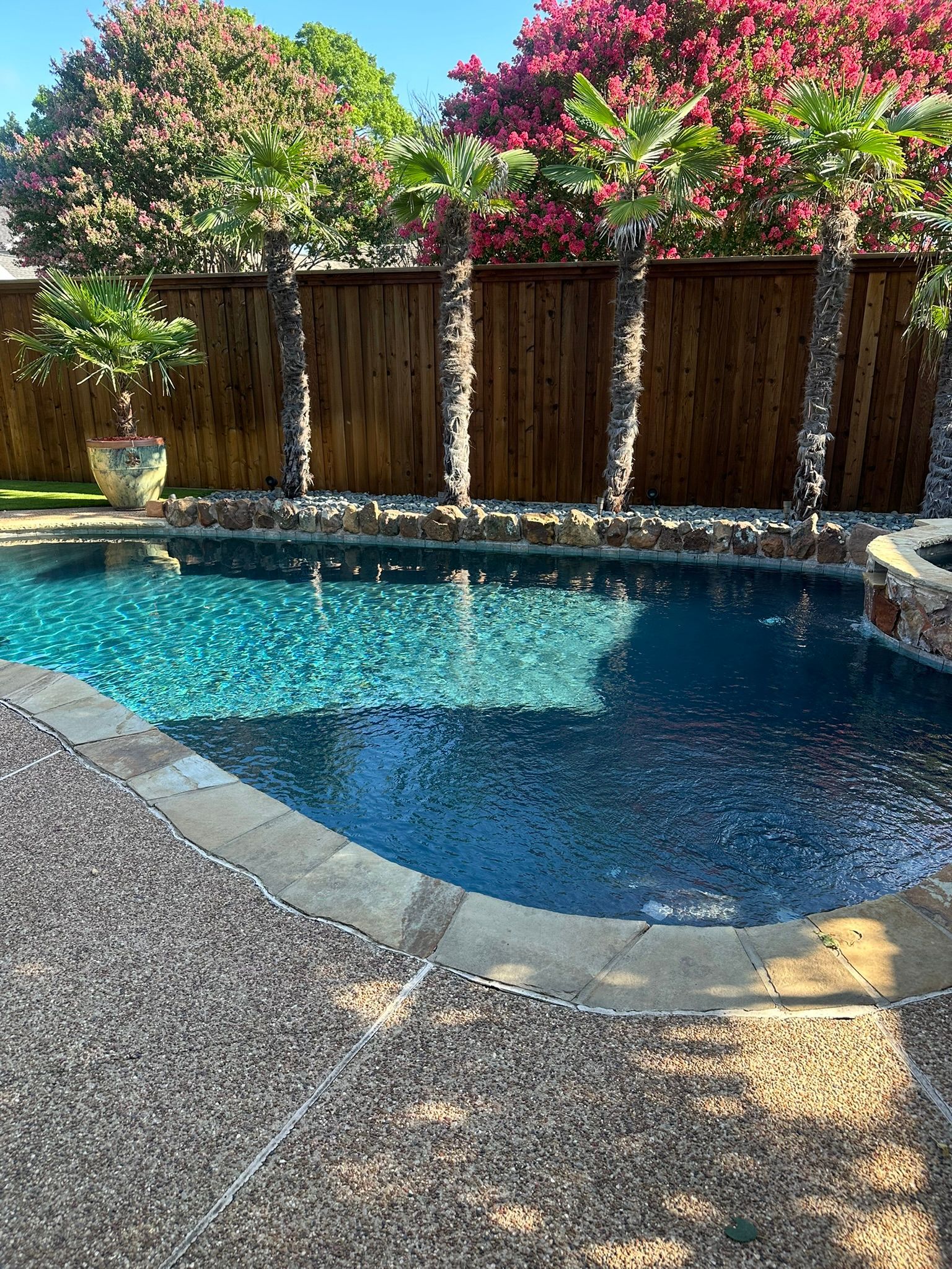 Pool with dark blue water and stone and wood fence, surrounded by a gravel patio.