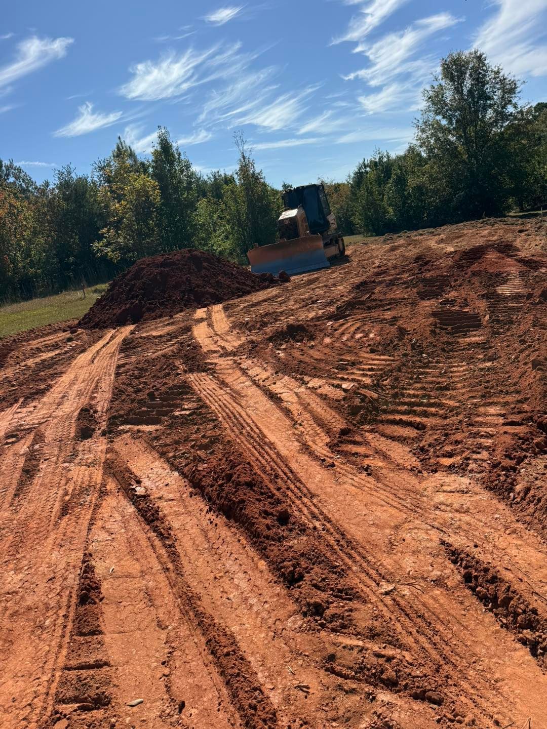 Bulldozer on a dirt hill, leaving tire tracks in red clay under a blue sky.