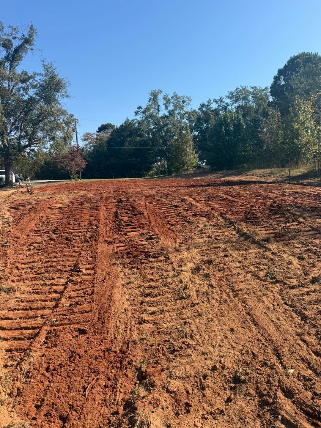 A cleared, red-dirt lot with tire tracks, trees in the background, under a blue sky.