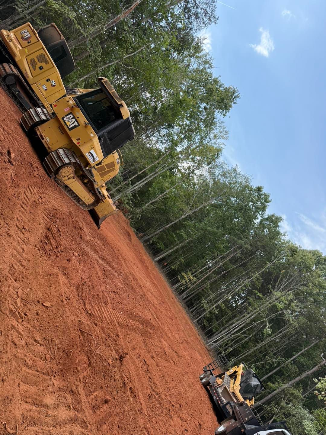 A yellow bulldozer grading red soil in a clearing with trees under a blue sky.