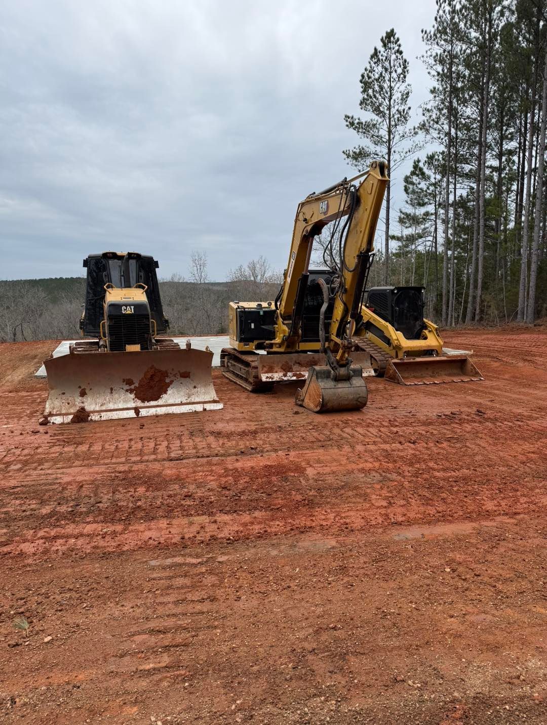 Three yellow construction vehicles on red dirt in a clearing, trees in background.