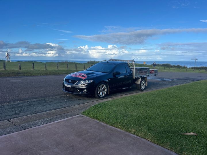 Black pickup truck with a trailer parked on a roadside near green grass and open water under a blue sky — P & D Garage Doors in Taree, NSW