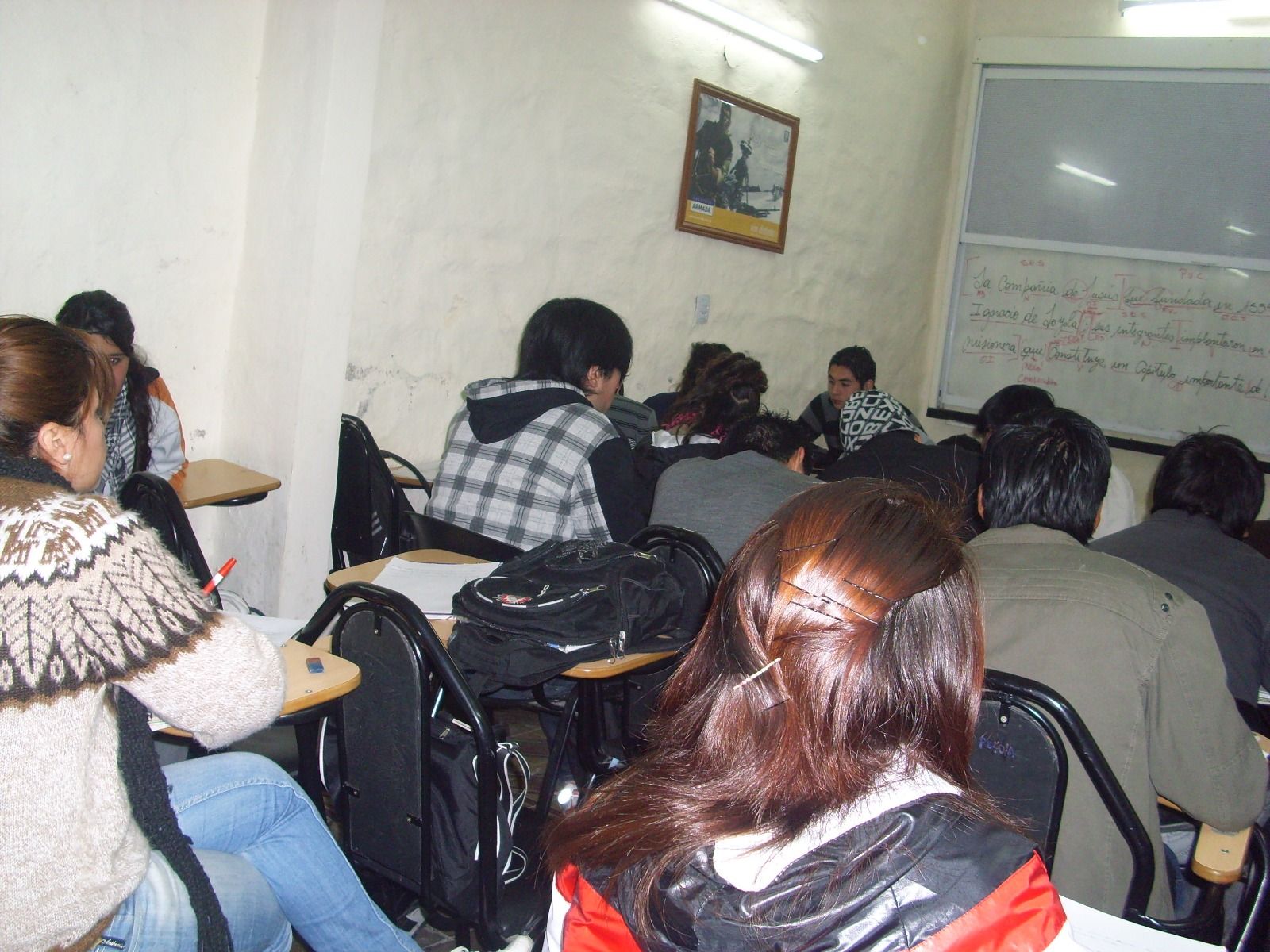Alumnos sentados en un aula, frente a una pizarra blanca con texto y una imagen en la pared.