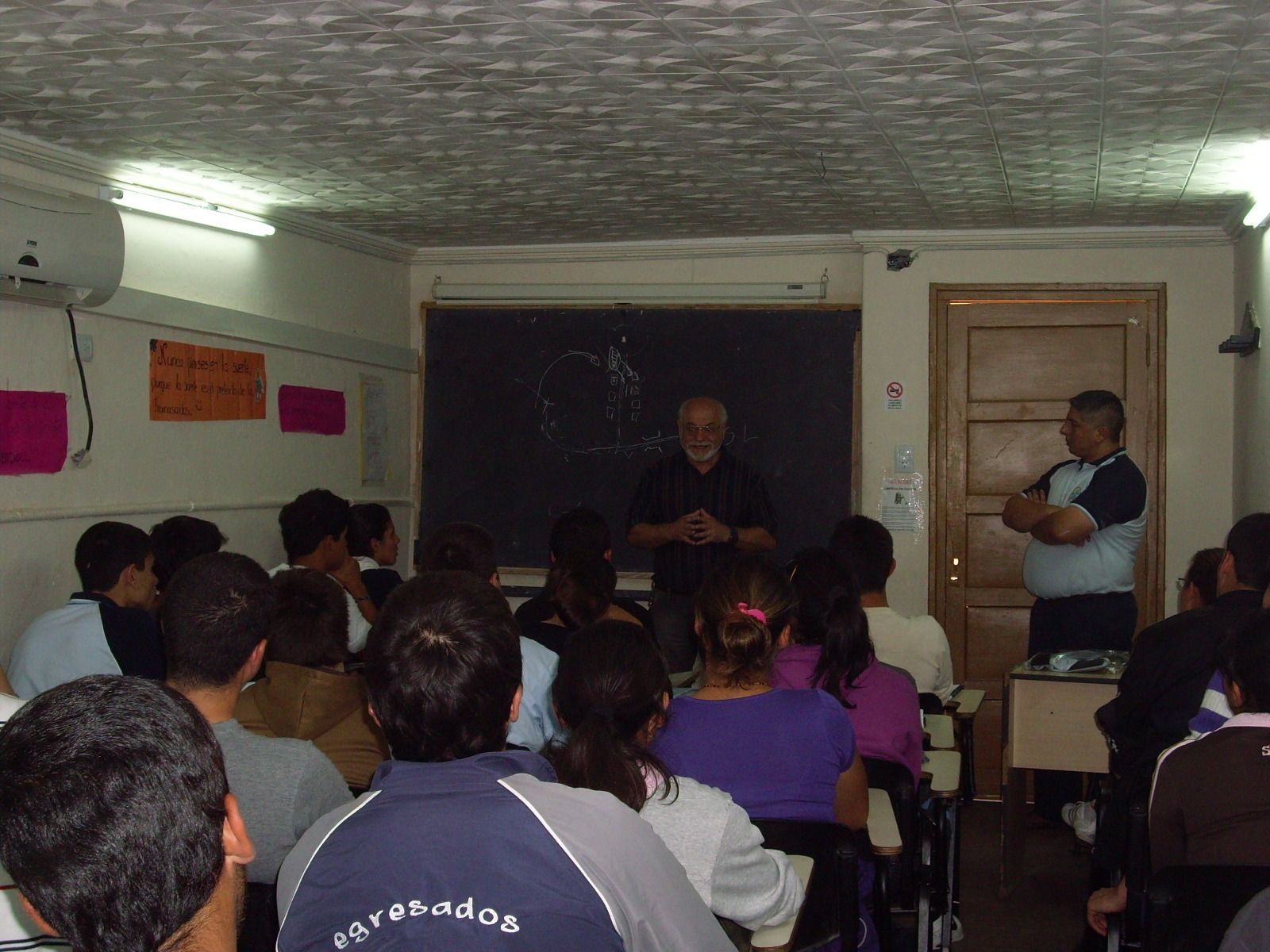 Aula pequeña con alumnos sentados frente a dos personas situadas al frente, cerca de una pizarra.