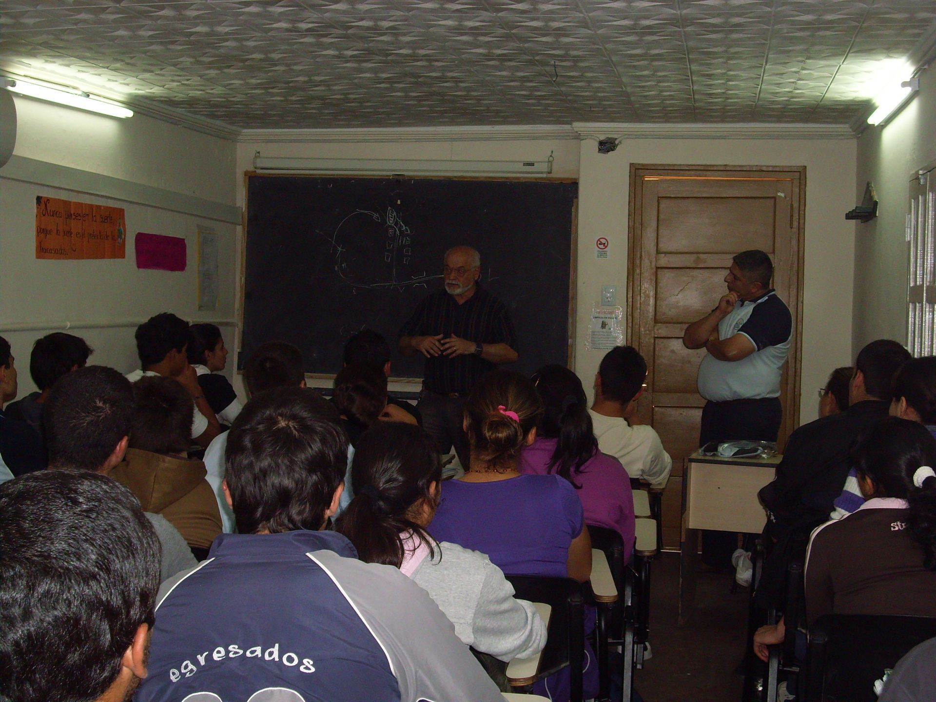 Clase magistral con los alumnos sentados frente al presentador, cerca de una pizarra y una puerta.