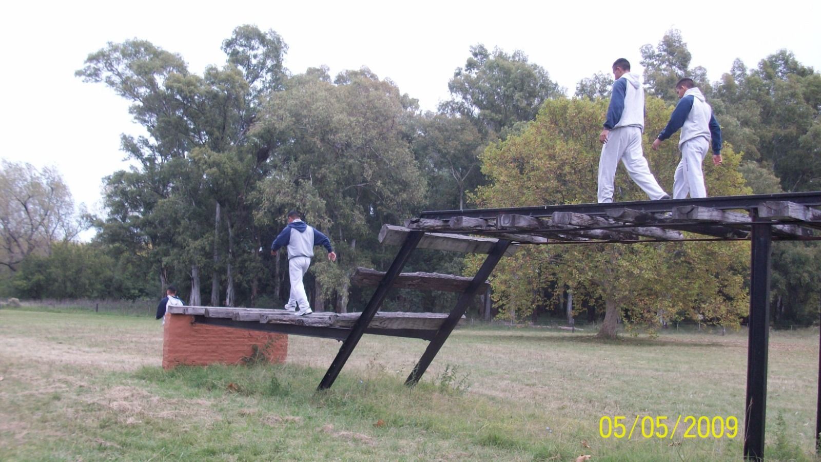 Tres personas con uniformes blancos en una pista de obstáculos de madera en un parque con césped.