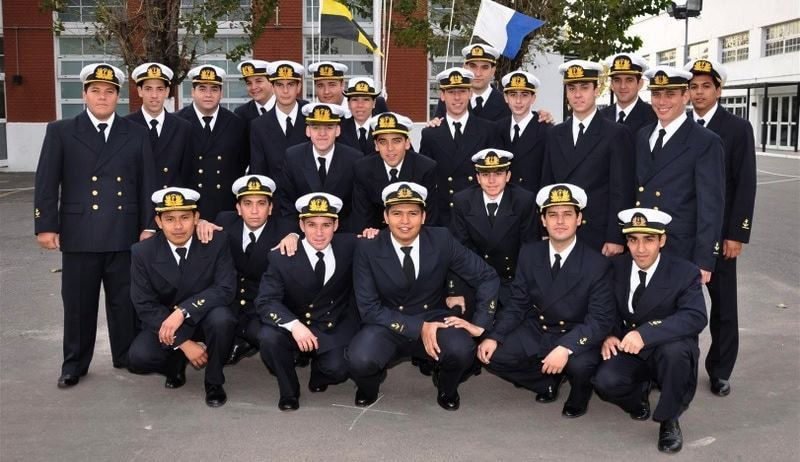 Grupo de oficiales navales con uniformes de la marina, algunos arrodillados, posando frente a un edificio con banderas.