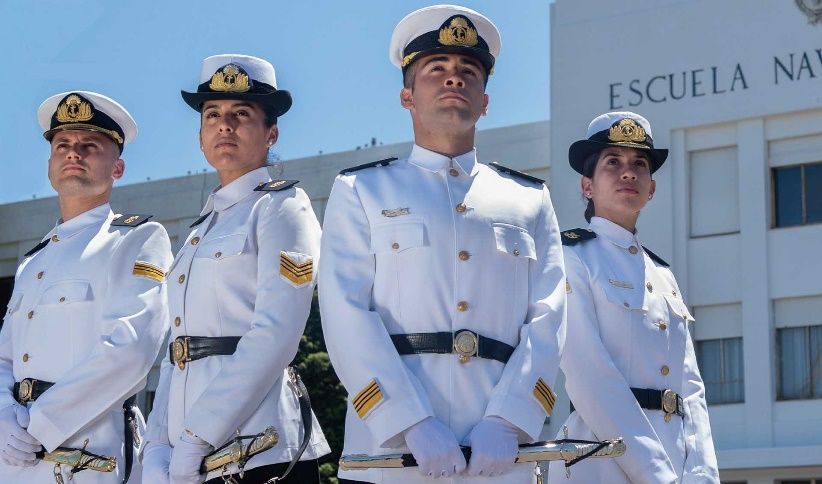 Cuatro oficiales navales con uniformes blancos se encuentran frente a un edificio etiquetado 