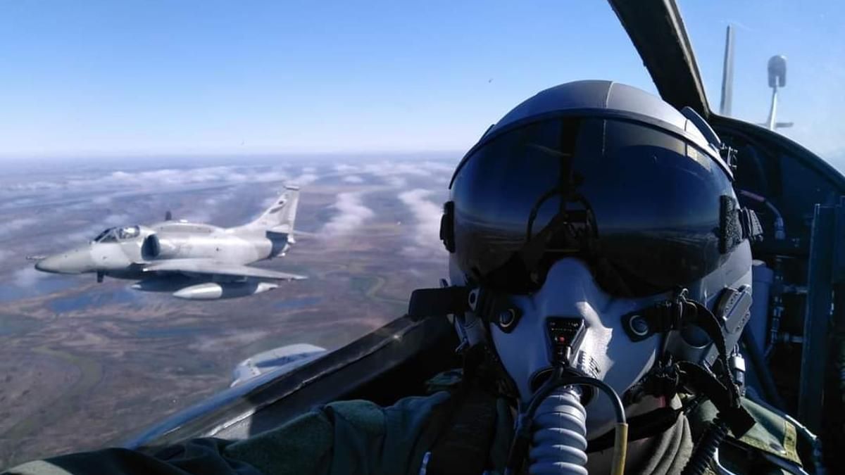 Piloto en cabina, con un avión a reacción volando cerca, contra un cielo azul.
