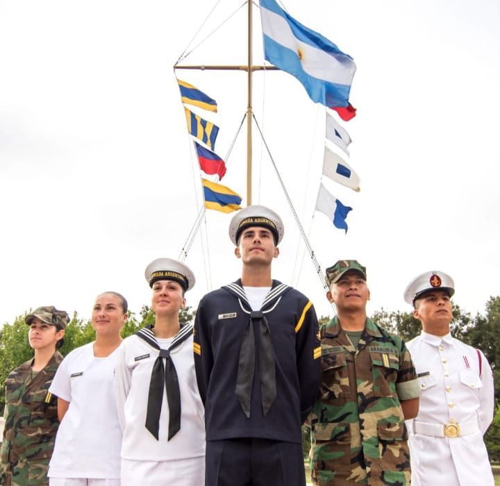 Cinco cadetes uniformados permanecen firmes bajo banderas sobre un fondo blanco.