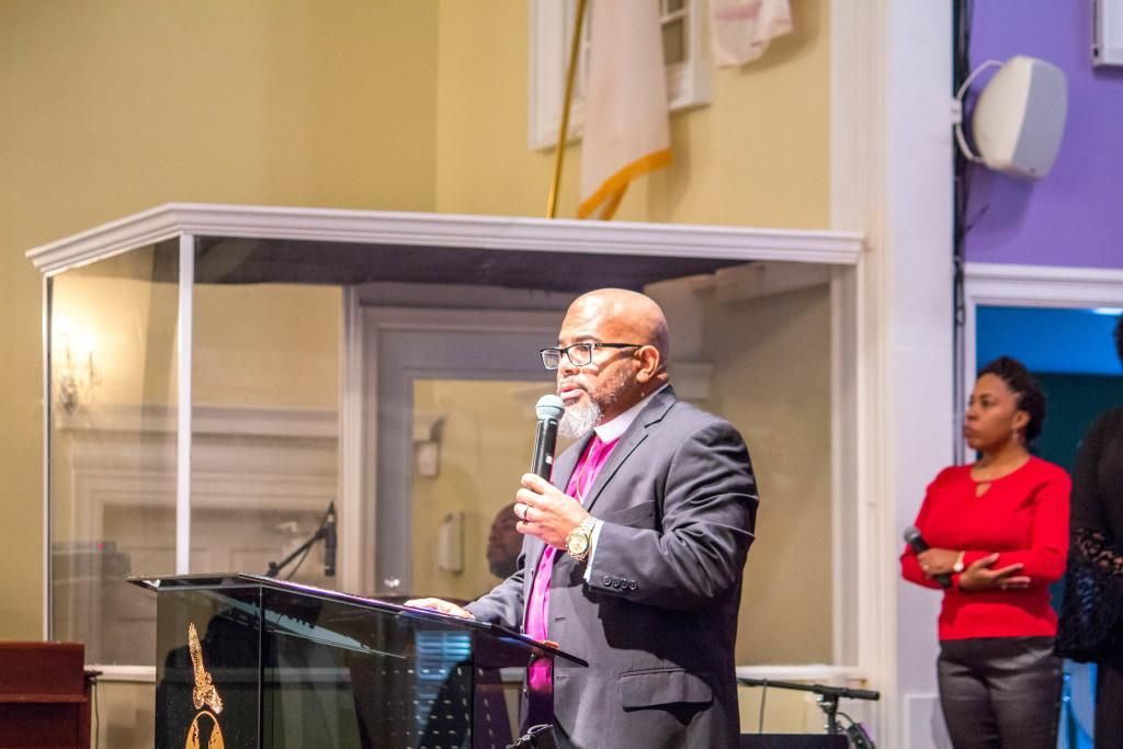 Man speaking at a podium in a church, holding a microphone. Other people stand nearby.