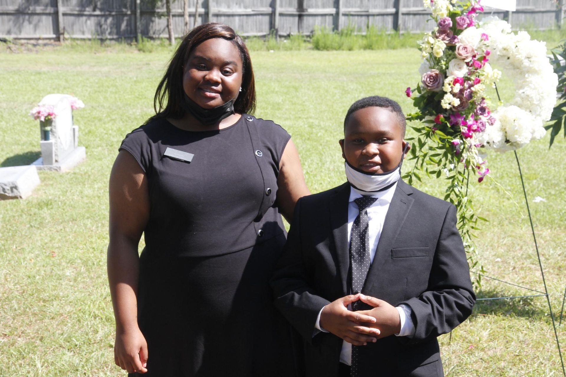 Woman and boy standing outside near flowers, both wearing masks and formal attire.