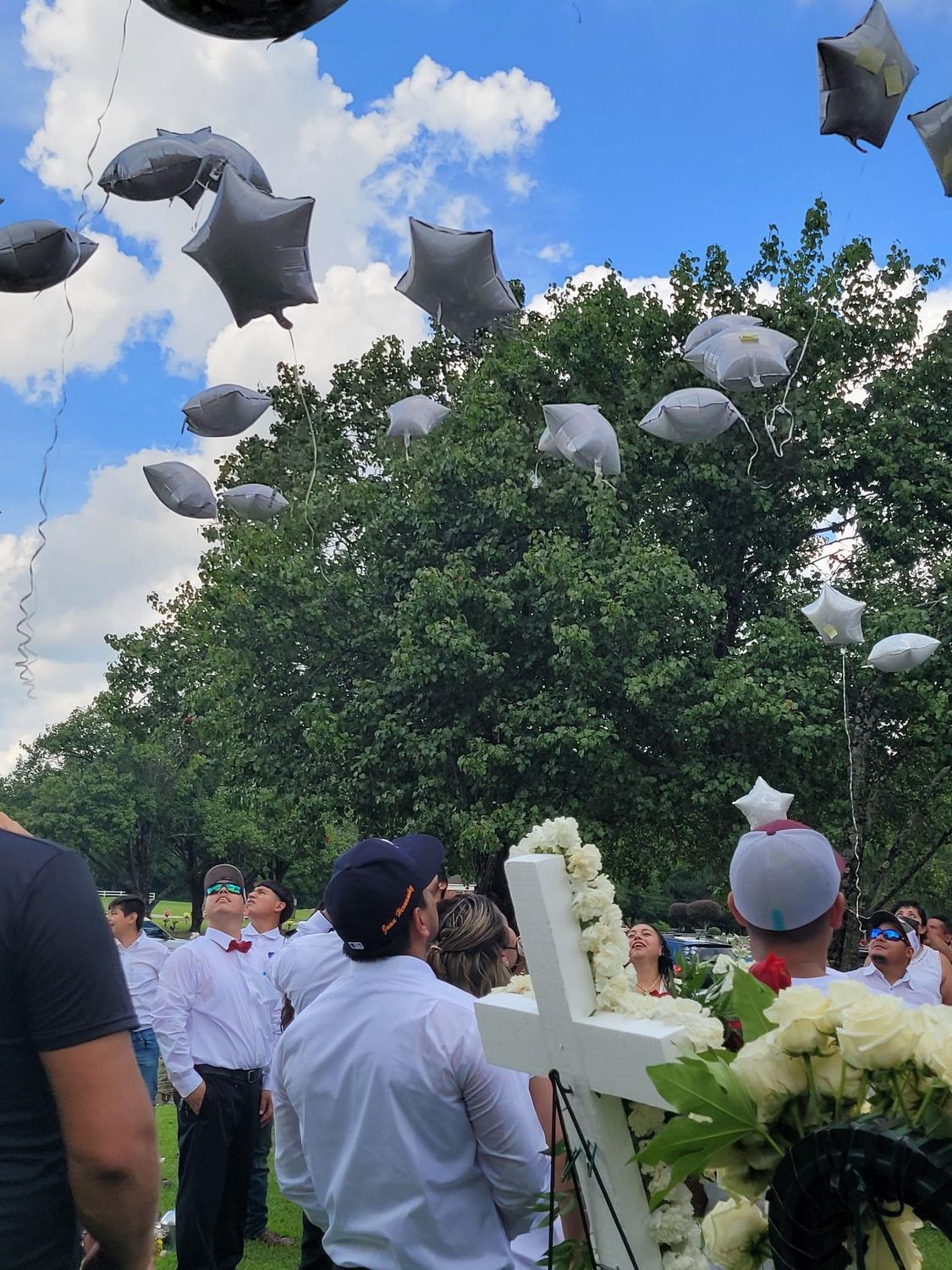 People releasing gray star-shaped balloons near a cross decorated with white flowers; outdoors, sunny day.