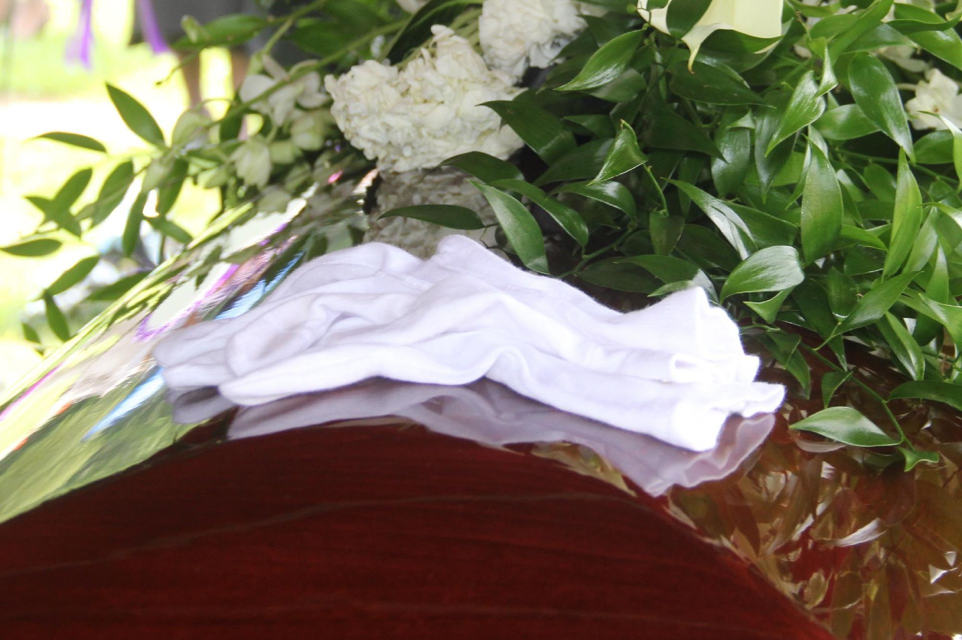 White cloth atop a polished wooden casket, adorned with white flowers and green foliage.
