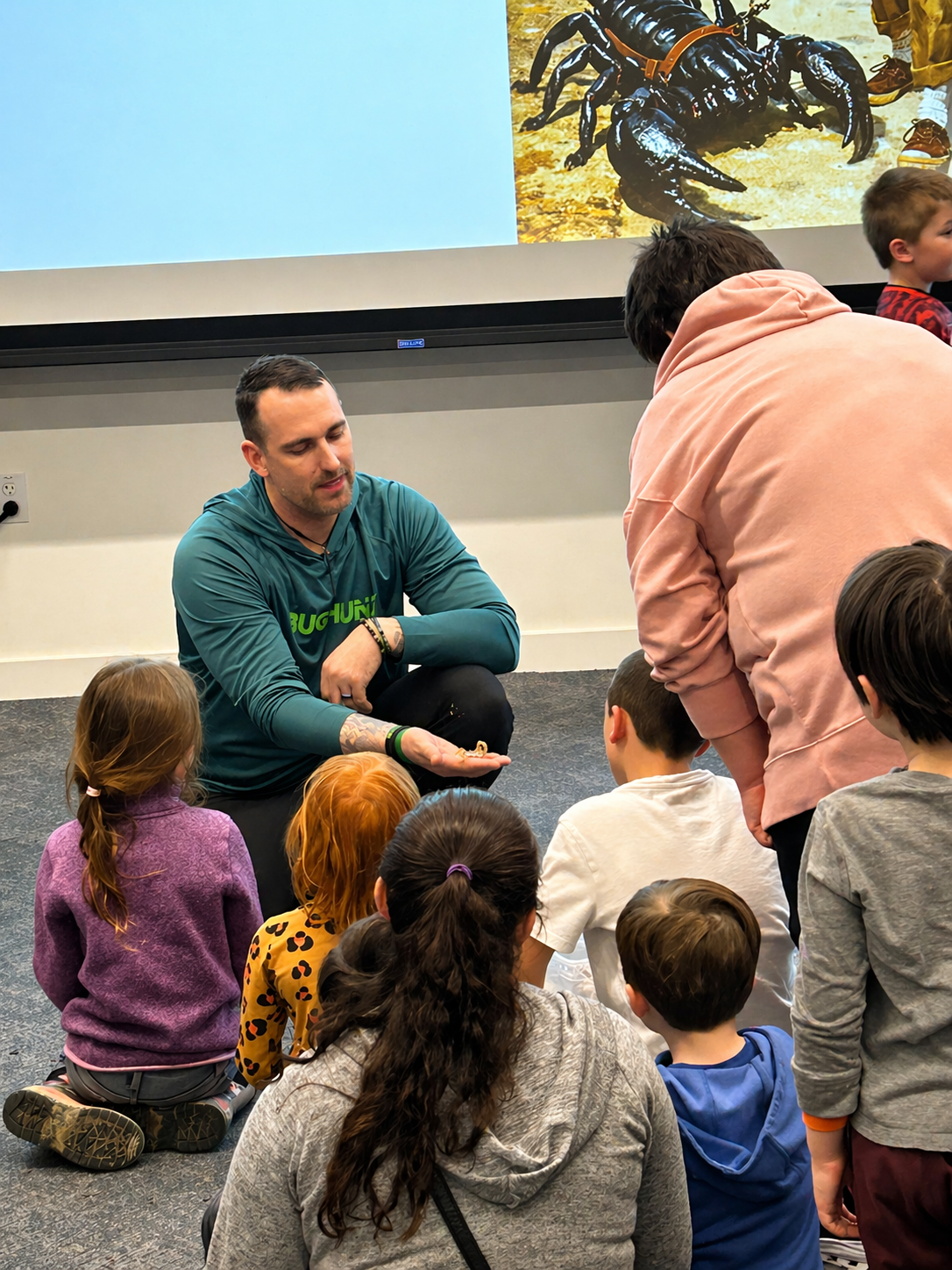 Speaker kneeling on the floor addressing a group of children in a classroom or assembly setting
