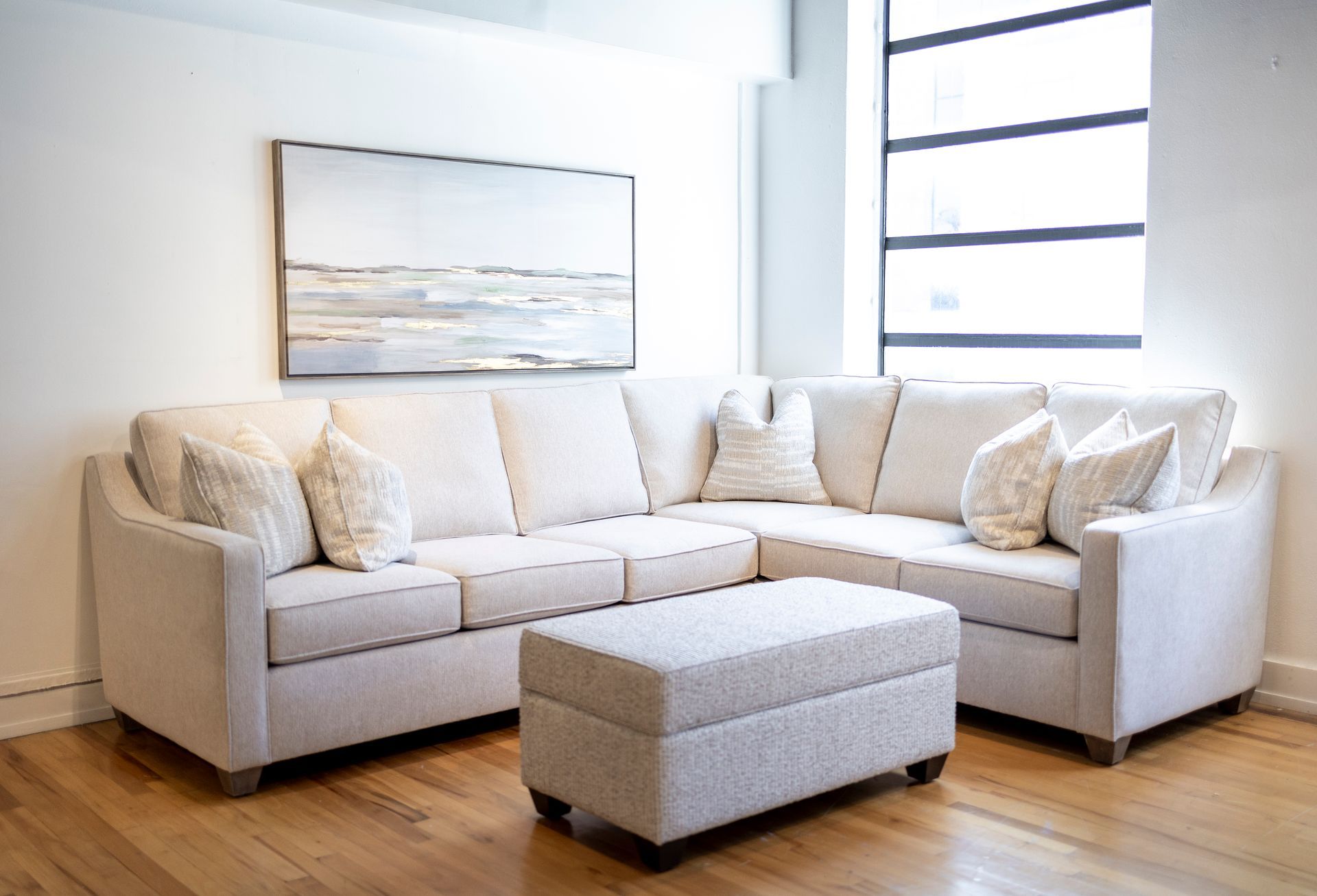 A living room with a sectional couch , ottoman , and painting on the wall.
