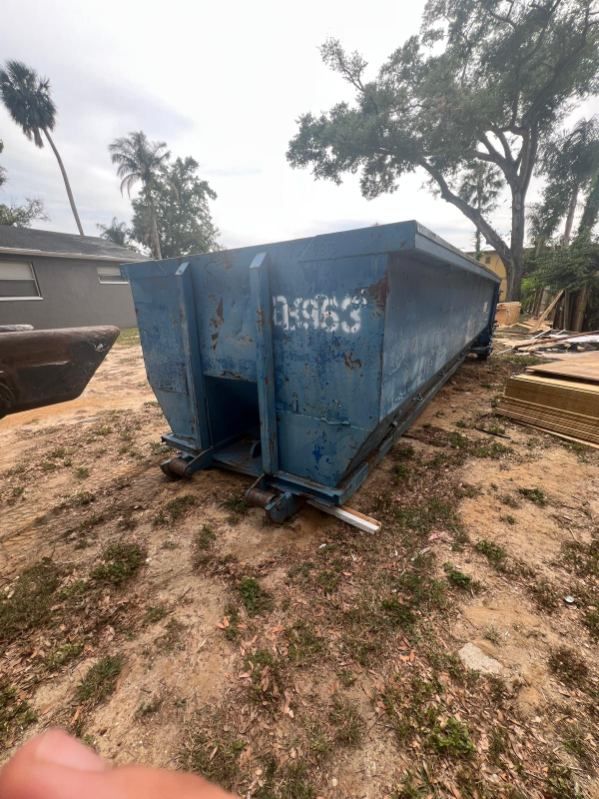 Blue metal dumpster on dirt in an outdoor yard, with trees and stacked lumber in the background.