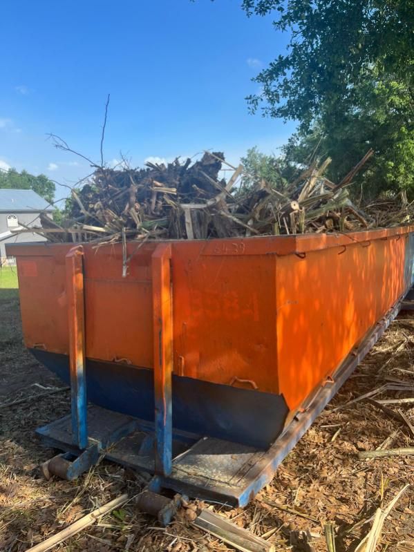 Orange metal dumpster full of yard debris, with blue supports, sitting outdoors in sunlight.